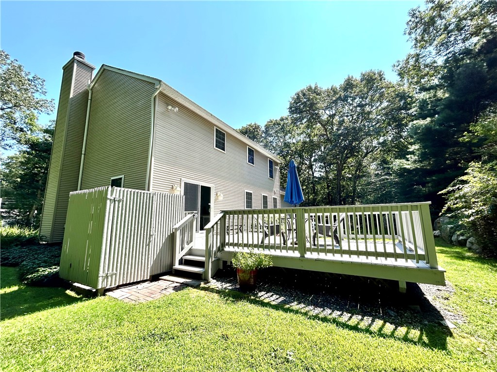Two-story house with a raised white deck, patio furniture, and a sunny lawn bordered by trees.
