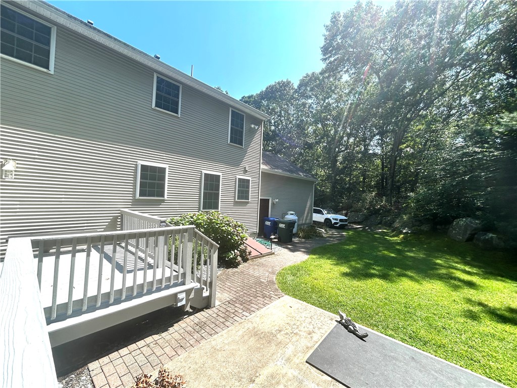 Two-story gray house with a raised deck, brick walkway, and sunny lawn bordered by mature trees.