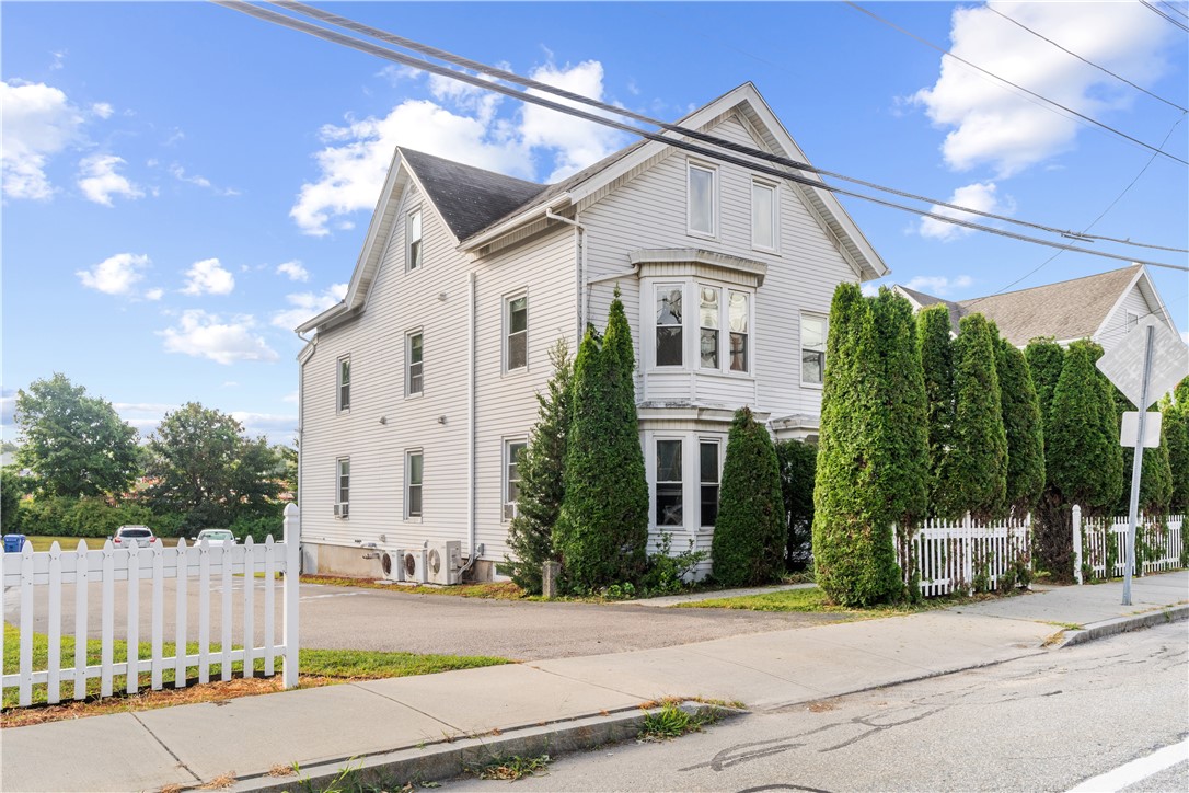 White multi-story house with bay windows, a picket fence, and tall evergreen shrubs.