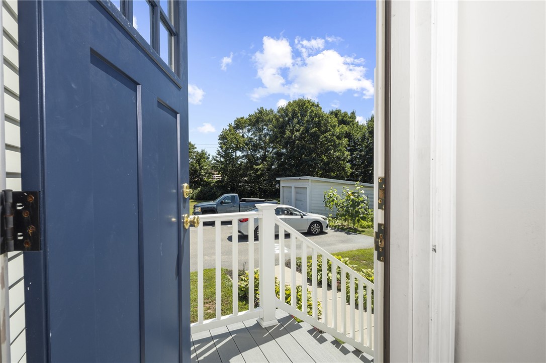 Open blue front door looking onto a small porch with white railings and a sunny parking area beyond.