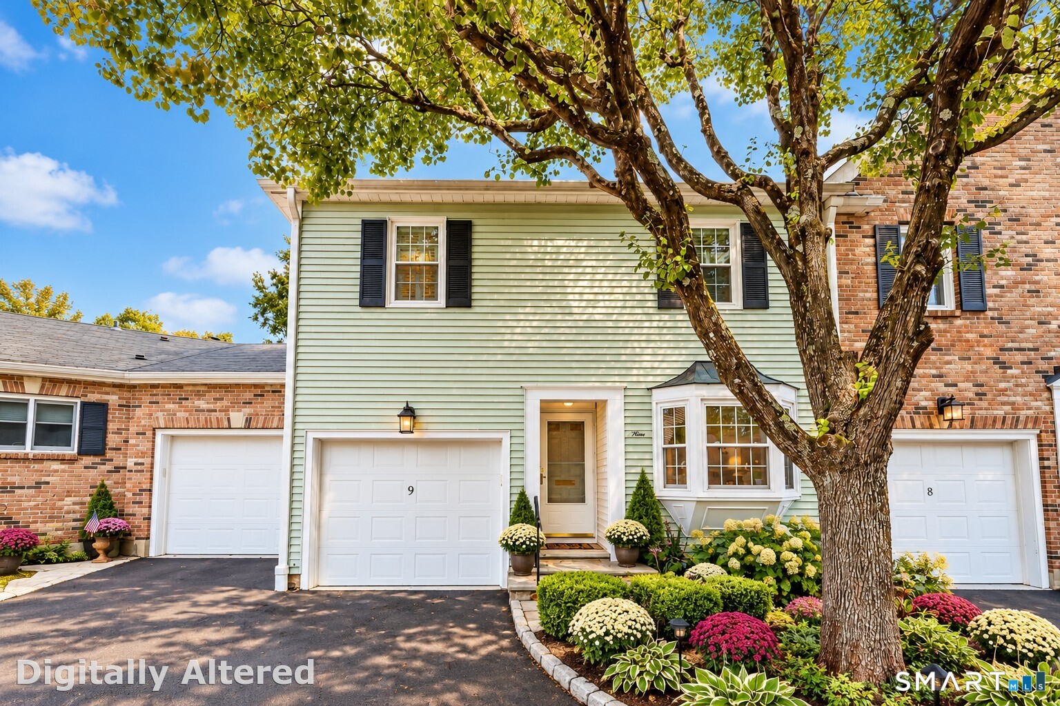 Unit 9 townhouse exterior with pale siding, brick neighbors, mature tree, and colorful landscaping.
