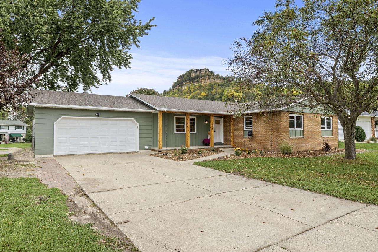 Ranch-style home with sage-green siding, brick facade, attached garage, and wooded bluff behind it.