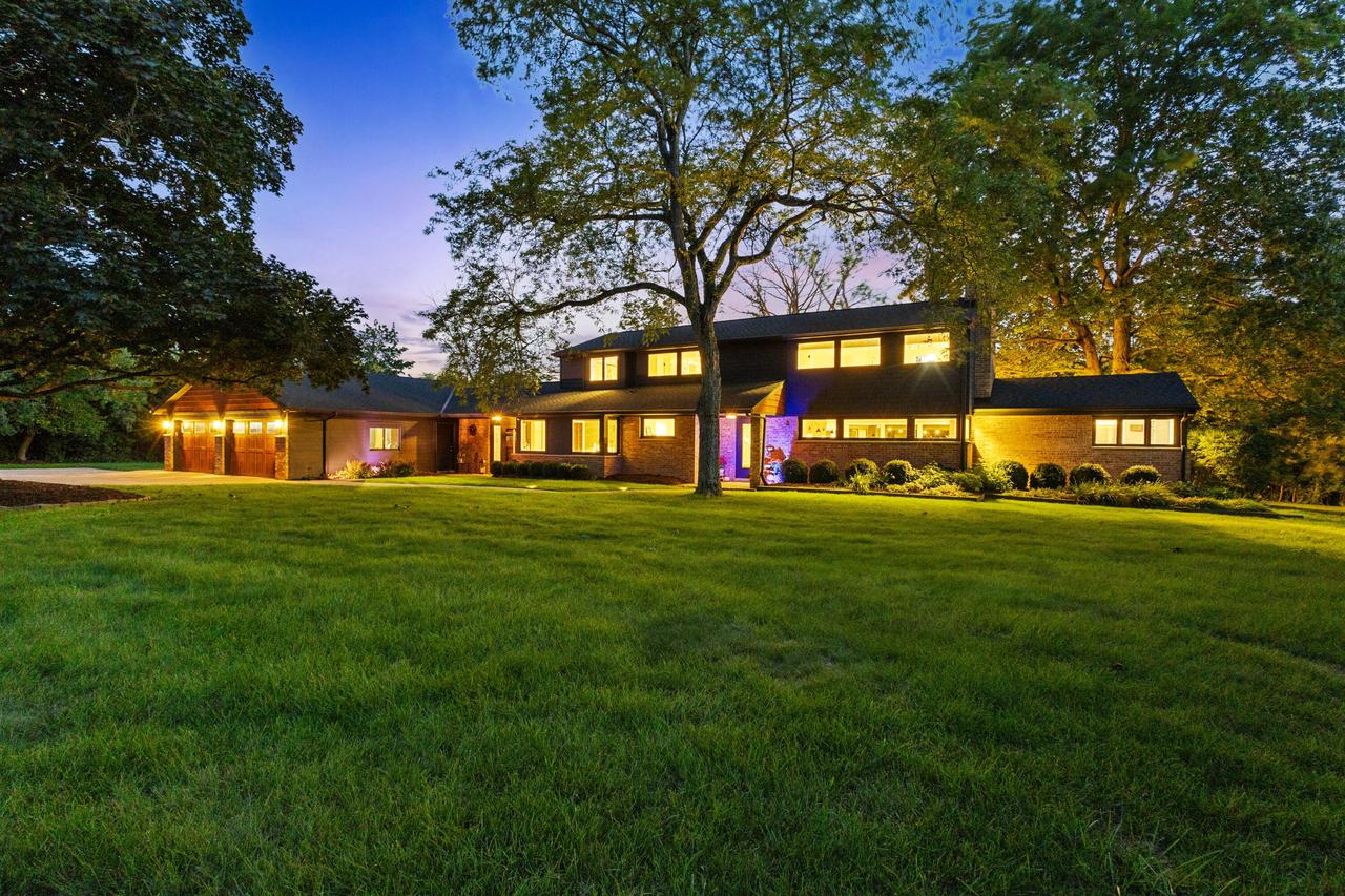 Brick home glowing at dusk beneath mature trees, with a broad front lawn.