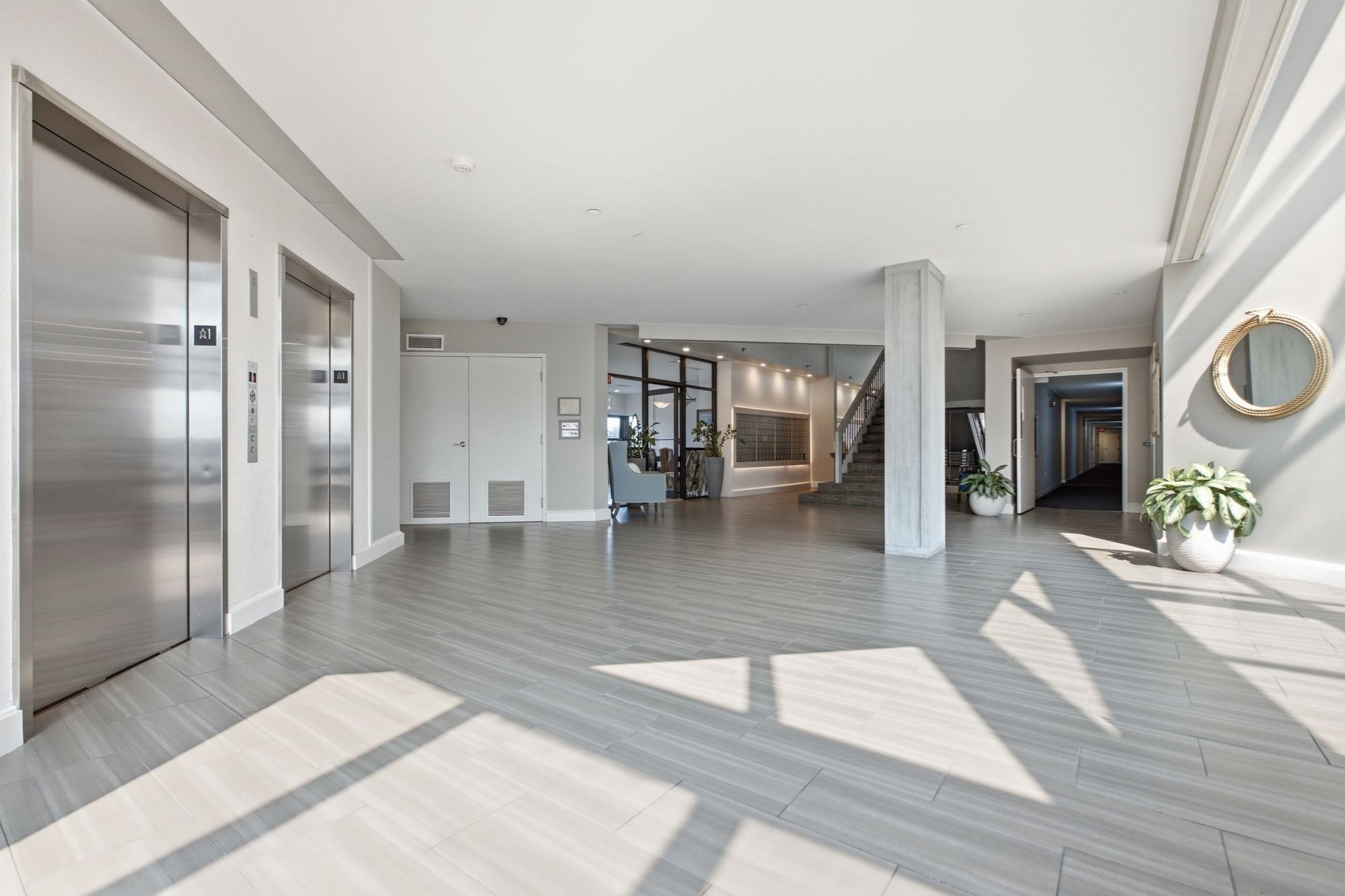Modern apartment lobby with stainless steel elevators, a staircase, and sunlit gray tile floors.