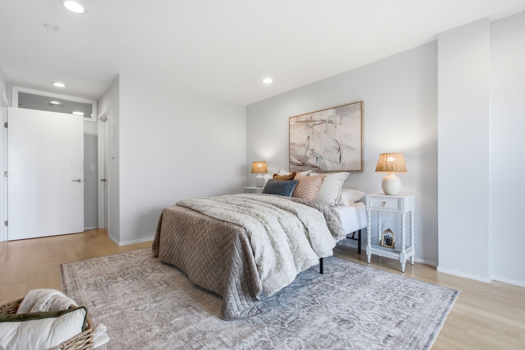 Staged bedroom with layered gray and white bedding, bedside lamps, and abstract artwork.