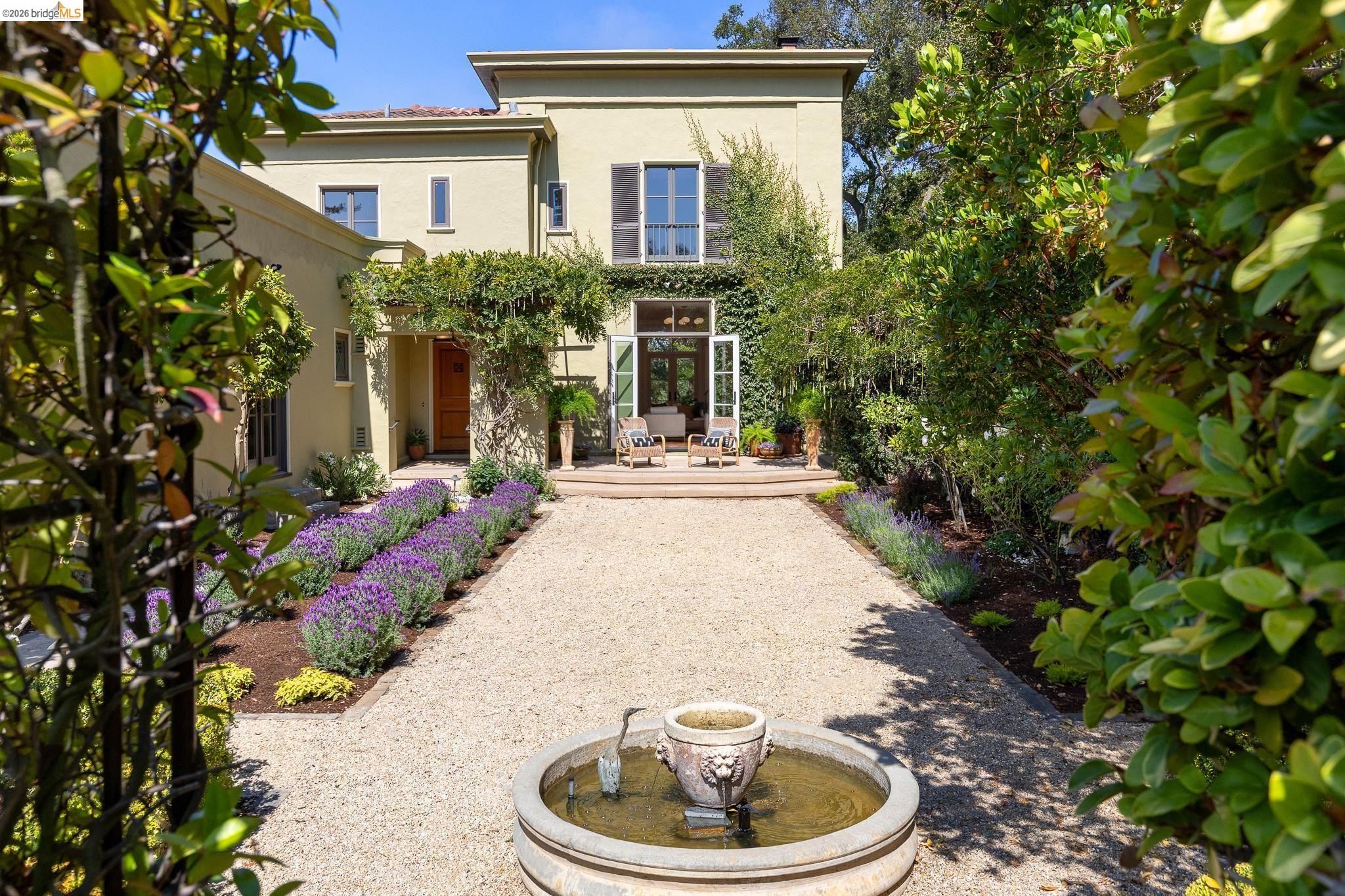 Cream two-story house beyond a gravel courtyard with a fountain, lavender beds, and a shaded patio.