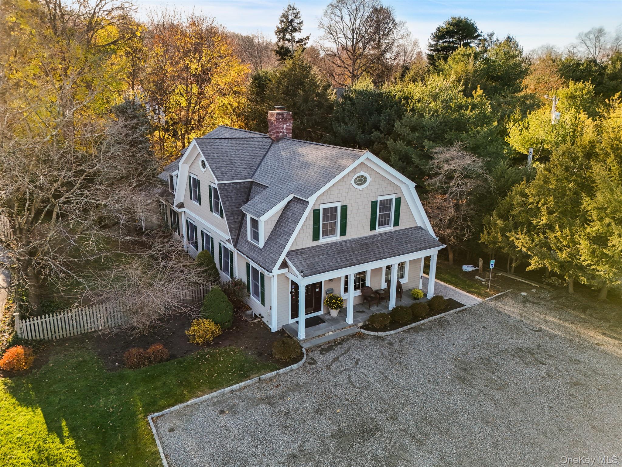 Two-story house with green shutters and a covered porch, surrounded by autumn trees and a gravel driveway.