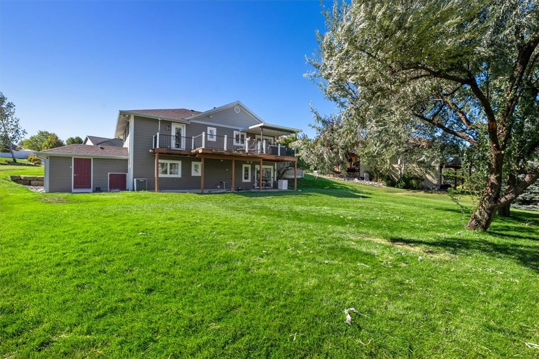 Gray two-story house with a raised deck overlooking a broad lawn and mature trees.