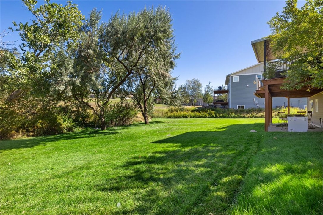 Backyard with a broad lawn, mature shade trees, and a covered patio beside a blue-sided house.