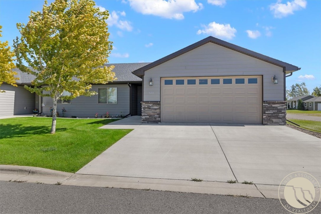 Gray single-story home with a two-car garage, wide driveway, stone accents, and a leafy front lawn.