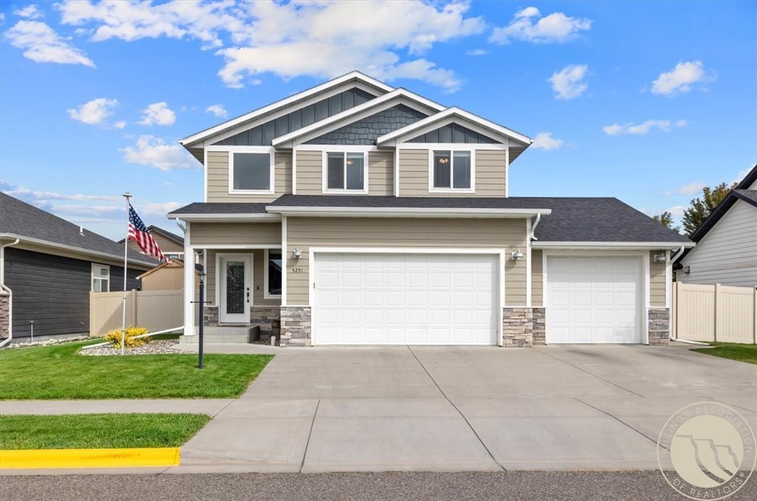 Two-story beige house with two white garage doors, a wide driveway, and an American flag.