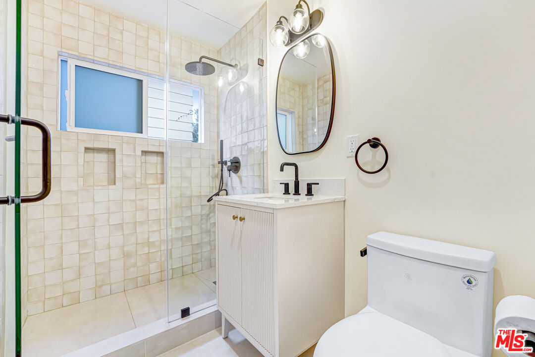Bathroom with a cream-tiled glass shower, white vanity, oval mirror, and toilet.