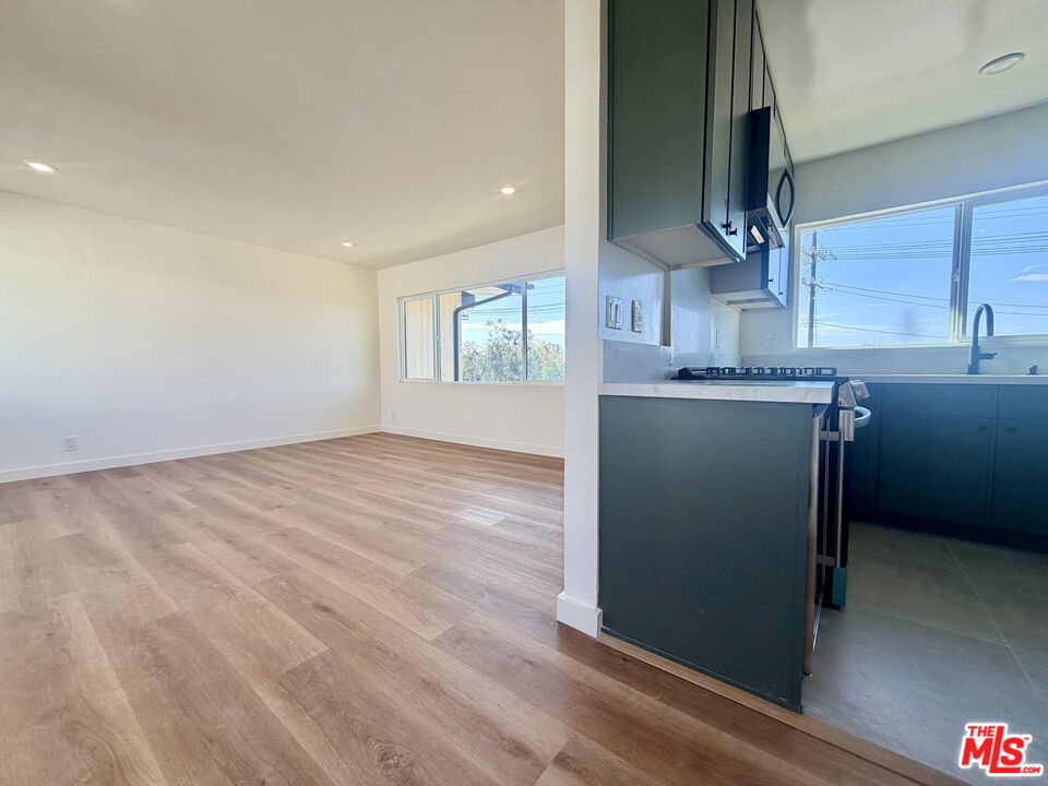 Vacant living area beside a dark-cabinet kitchen, with wood flooring and large windows.