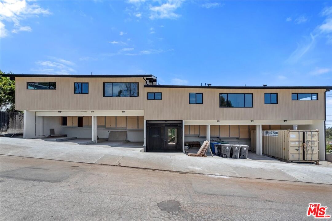 Modern two-story building with a flat roof, long rows of windows, and construction materials beside the driveway.