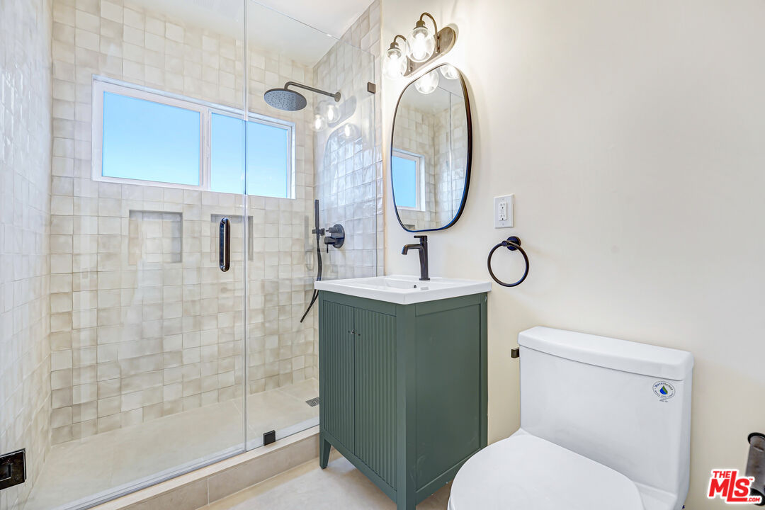 Bathroom with a tiled glass shower, green vanity, oval mirror, and white toilet.