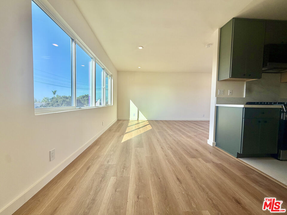 Vacant living and dining area with light wood floors, large windows, and dark kitchen cabinetry.