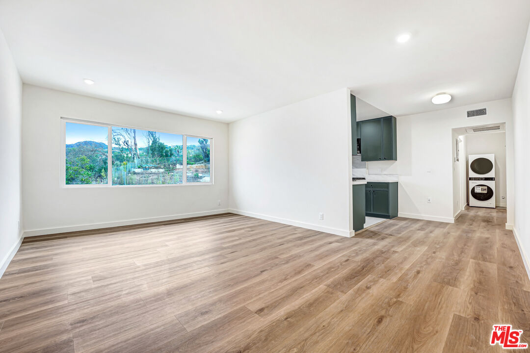 Vacant living area with wood floors, a wide window, dark-green kitchen cabinets, and a stacked washer and dryer.