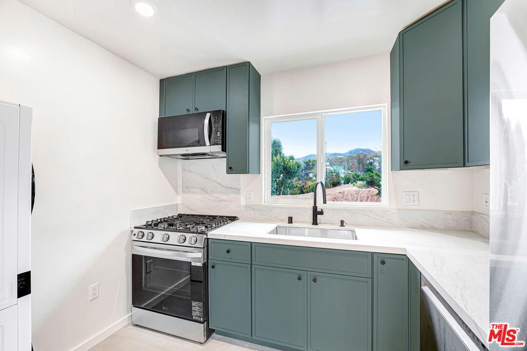 Kitchen with blue-green cabinets, marble counters, and a window overlooking trees and distant hills.