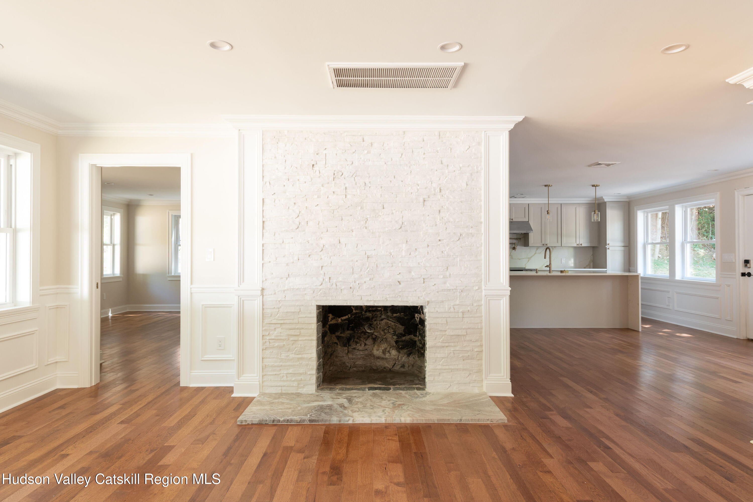 Living room with a white stone fireplace, hardwood floors, and the kitchen beyond.