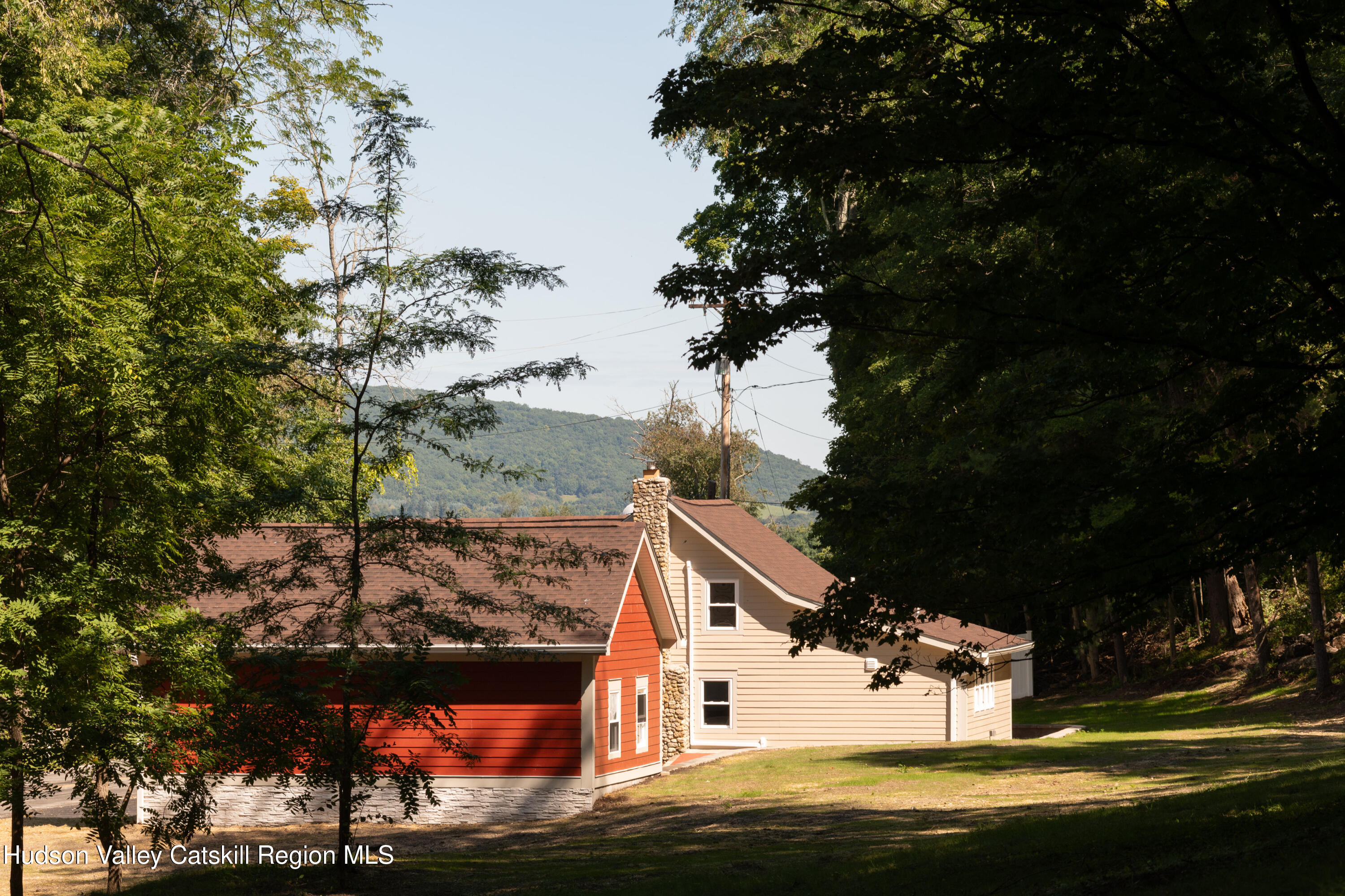 Red and beige house with a stone chimney among trees, with a forested mountain behind it.