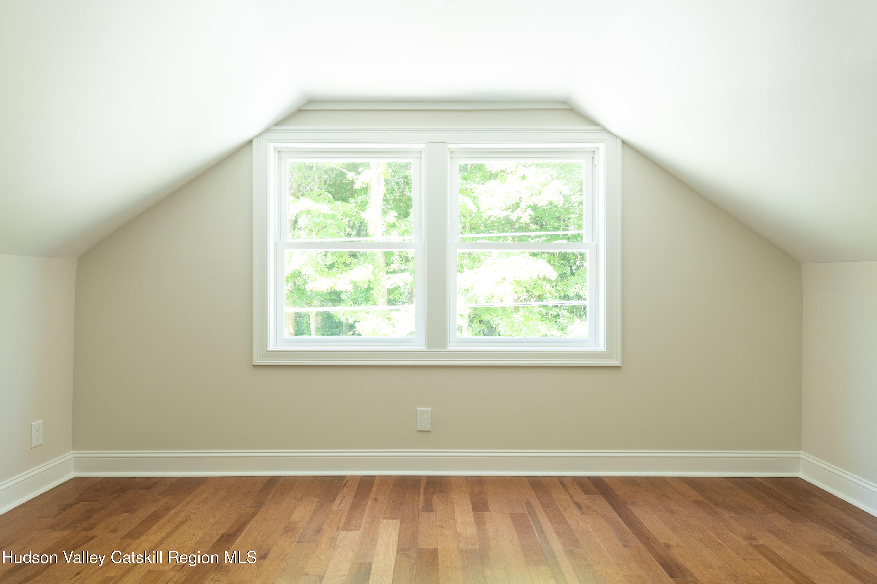 Empty upstairs room with sloped ceilings, hardwood floors, and a large double window overlooking trees.