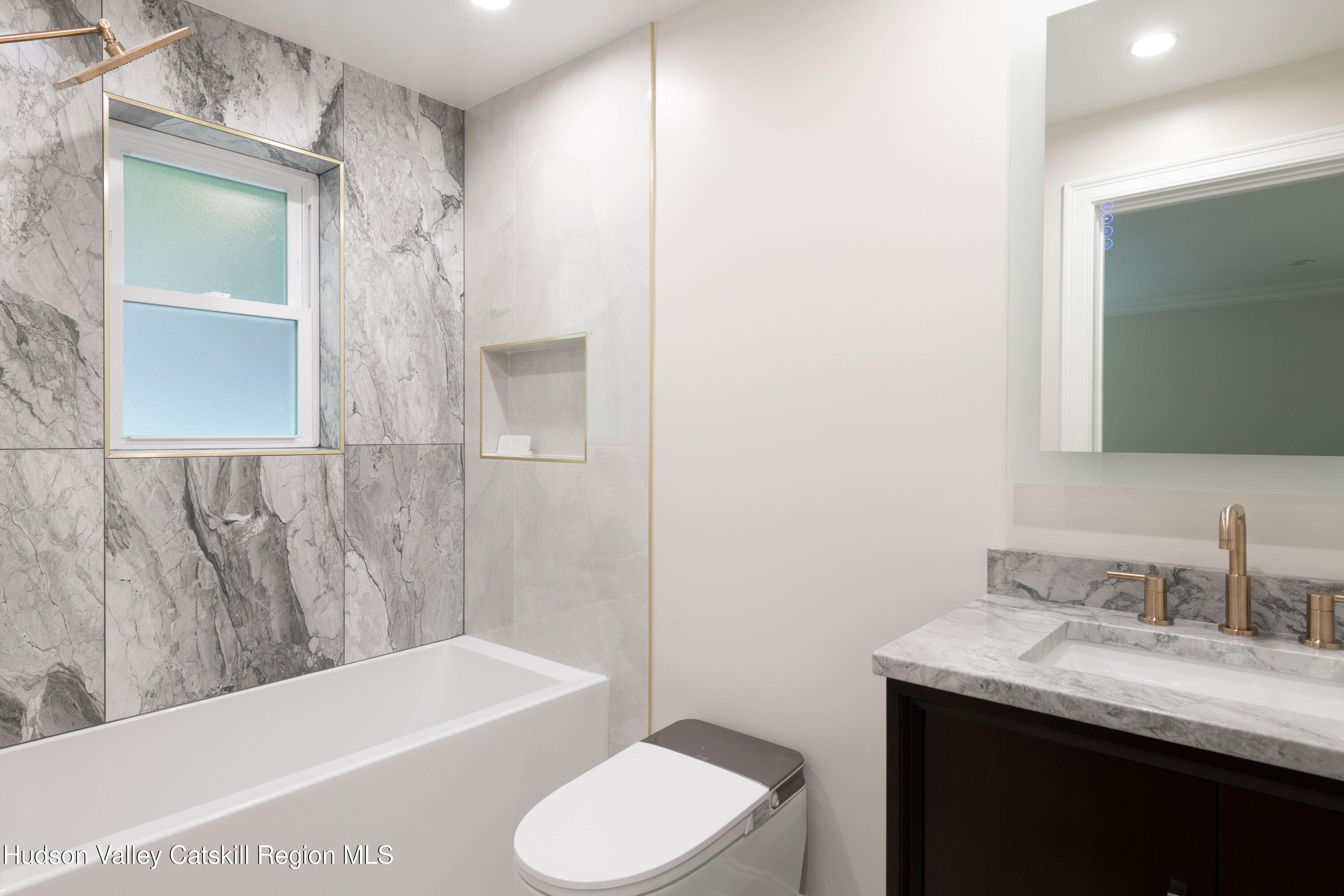 Bathroom with gray marble tub surround, frosted window, marble vanity, and brass fixtures.