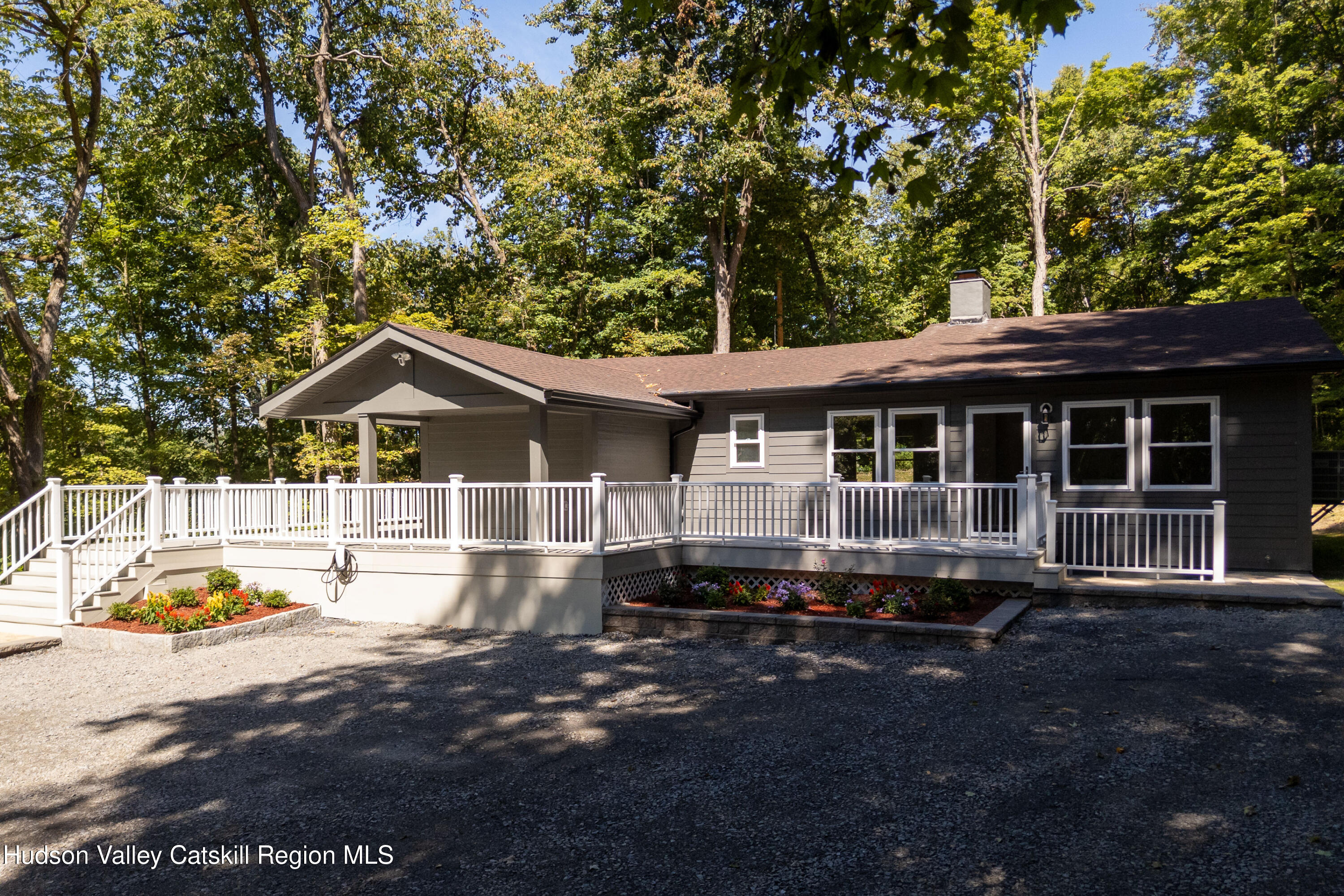 Gray single-story home with a large white-railed deck, flower beds, and a wooded backdrop.