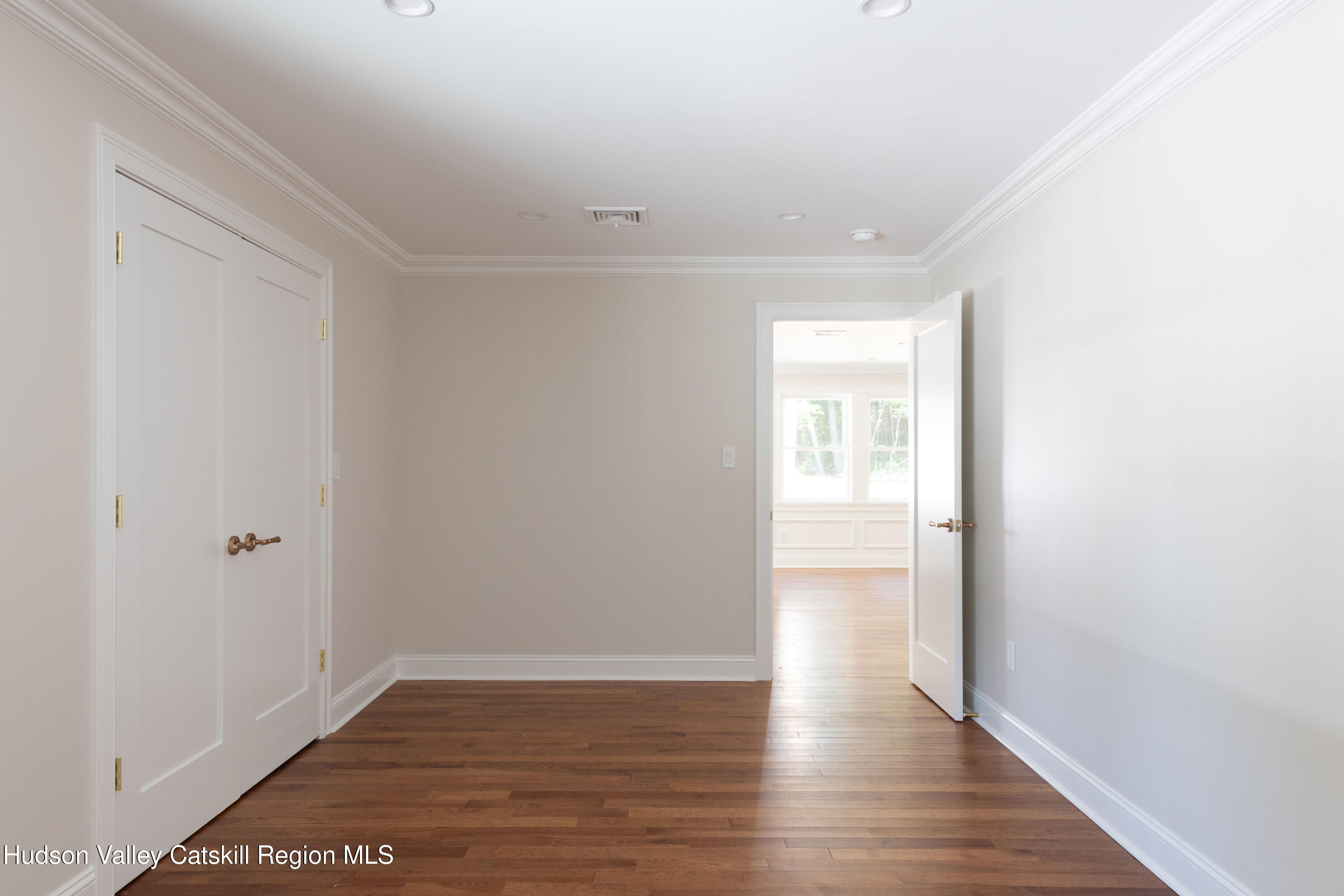 Empty room with dark hardwood floors and double closet doors, opening to a bright adjoining room.