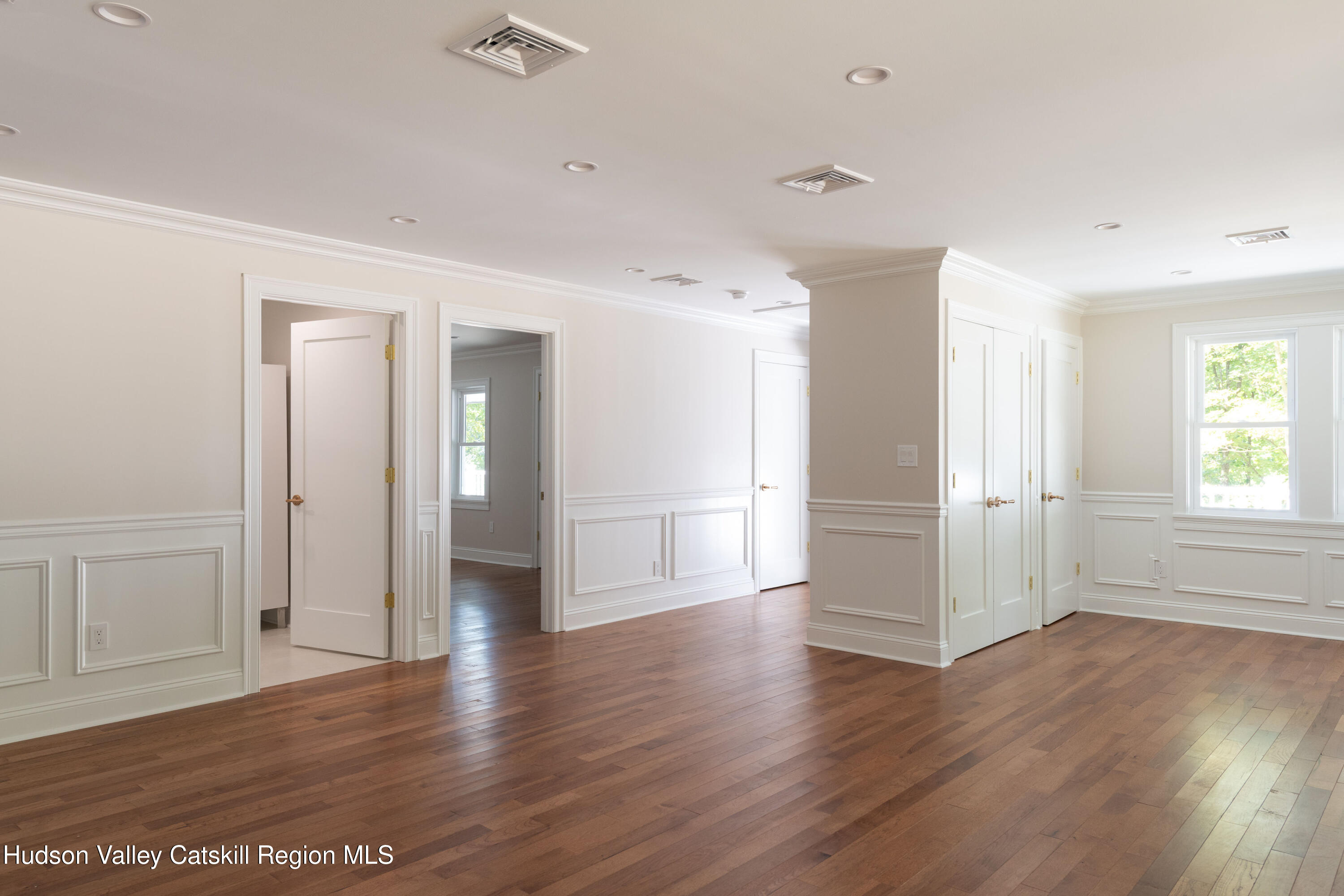 Empty room with hardwood floors, white paneled walls, multiple doorways, and a sunlit window.