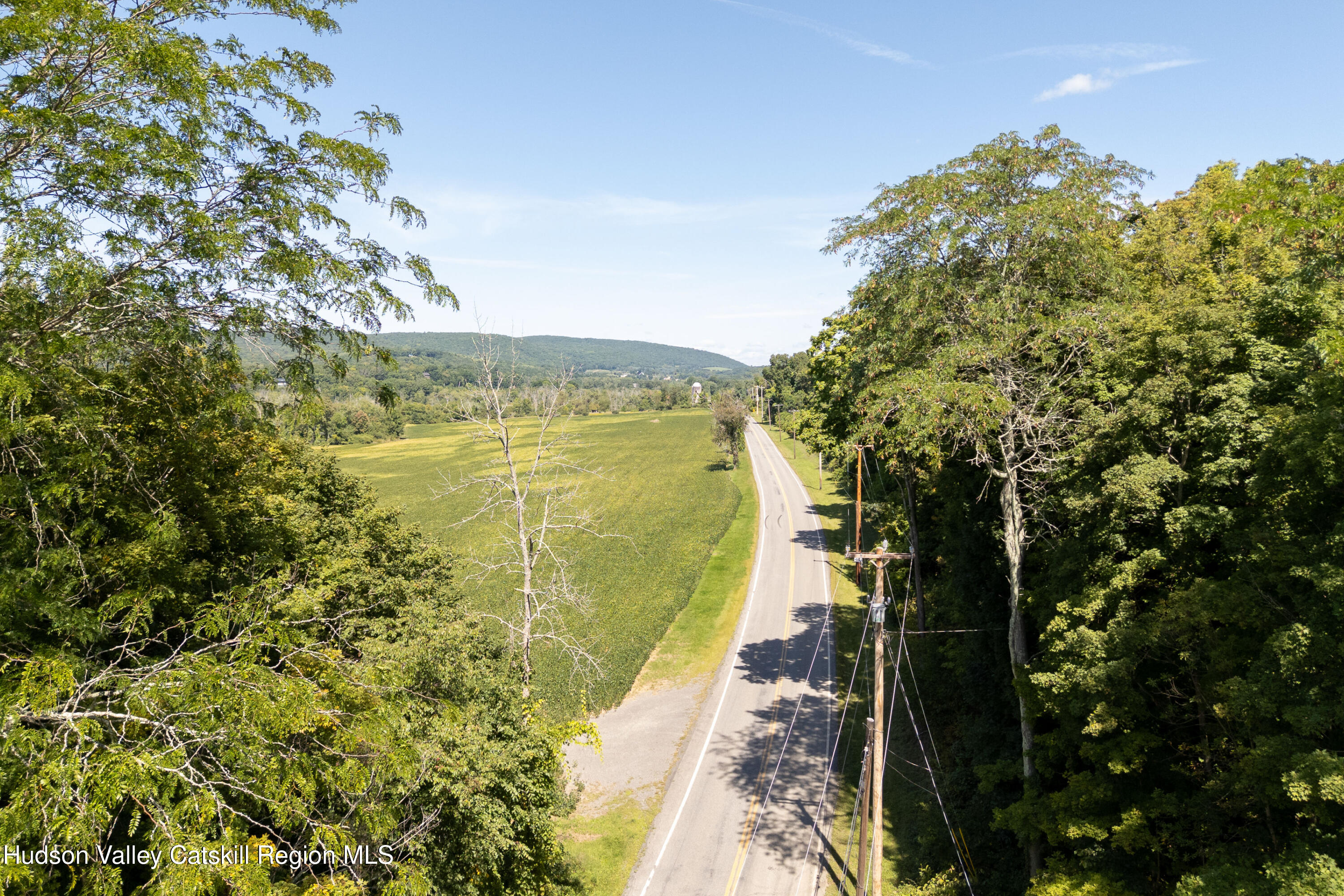Aerial view of a country road beside open fields and wooded hills.
