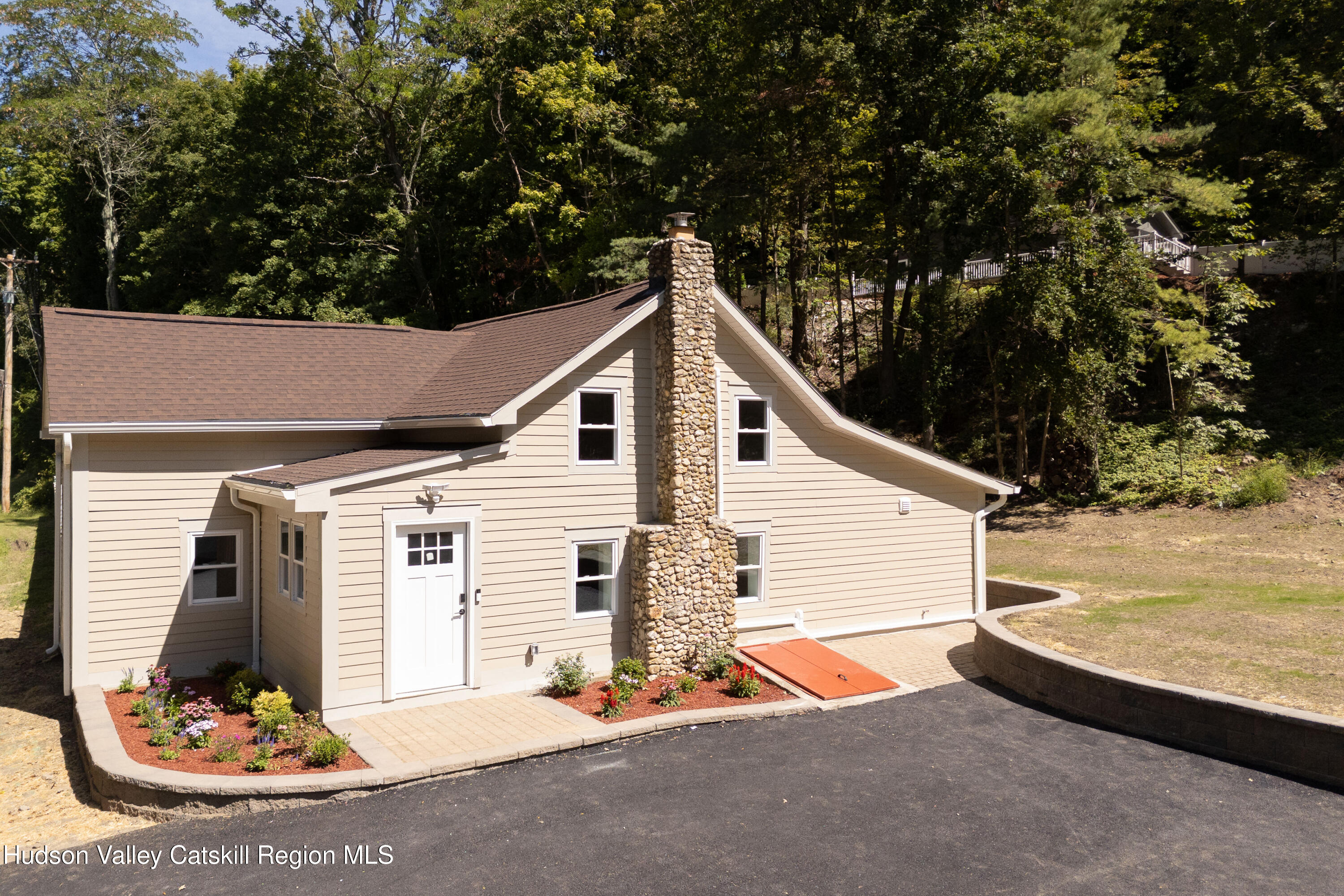 Beige-sided house with a tall stone chimney, flower beds, and a paved driveway among trees.