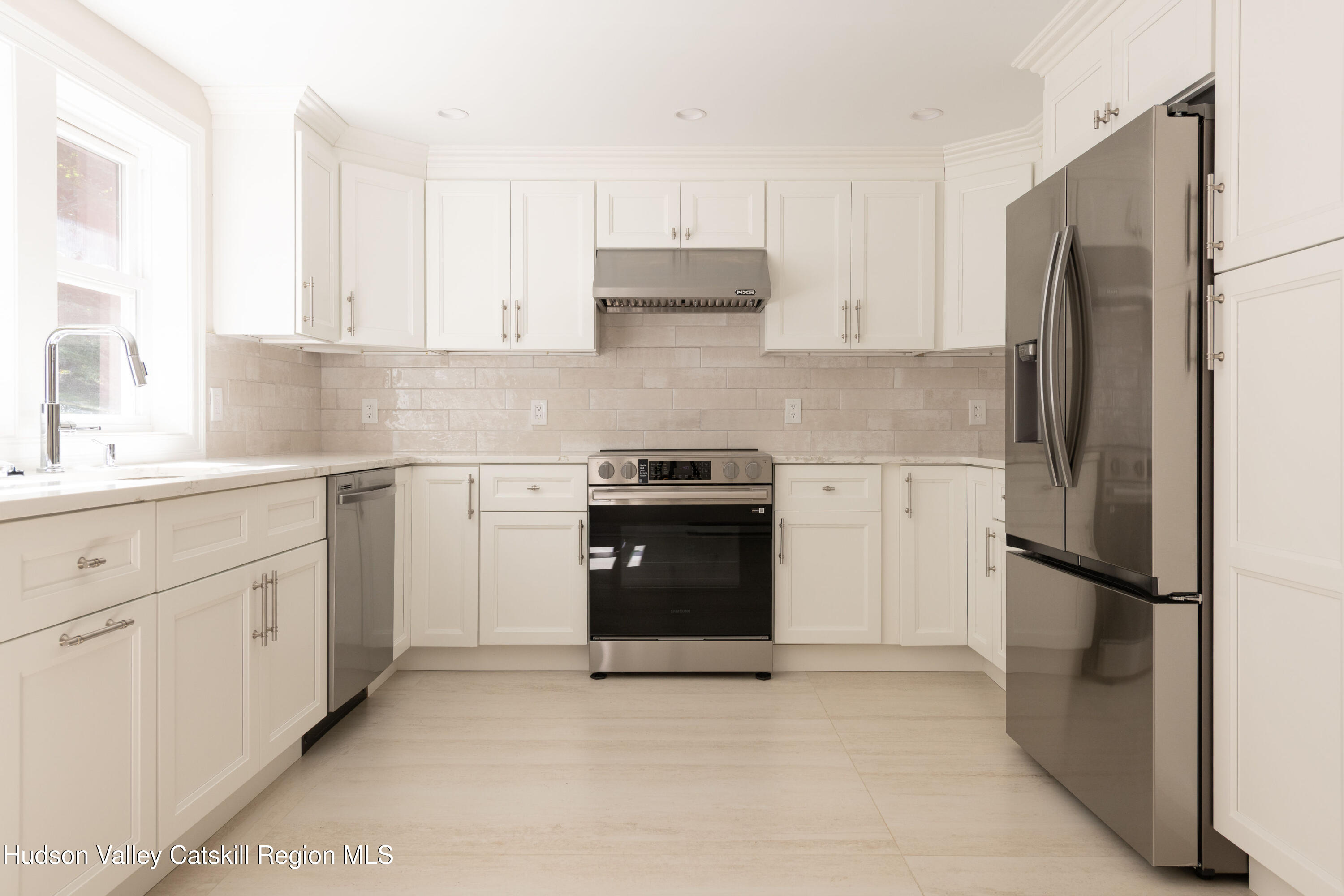 Bright white kitchen with stainless steel range and refrigerator, tile backsplash, and window over the sink.