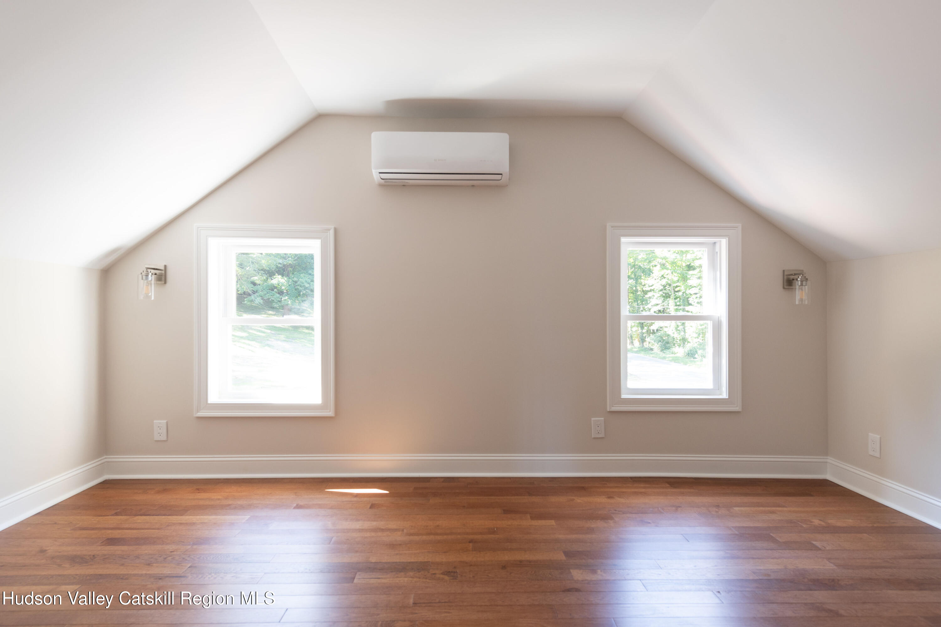 Empty room with a sloped ceiling, hardwood floors, two windows, and a wall-mounted mini-split.