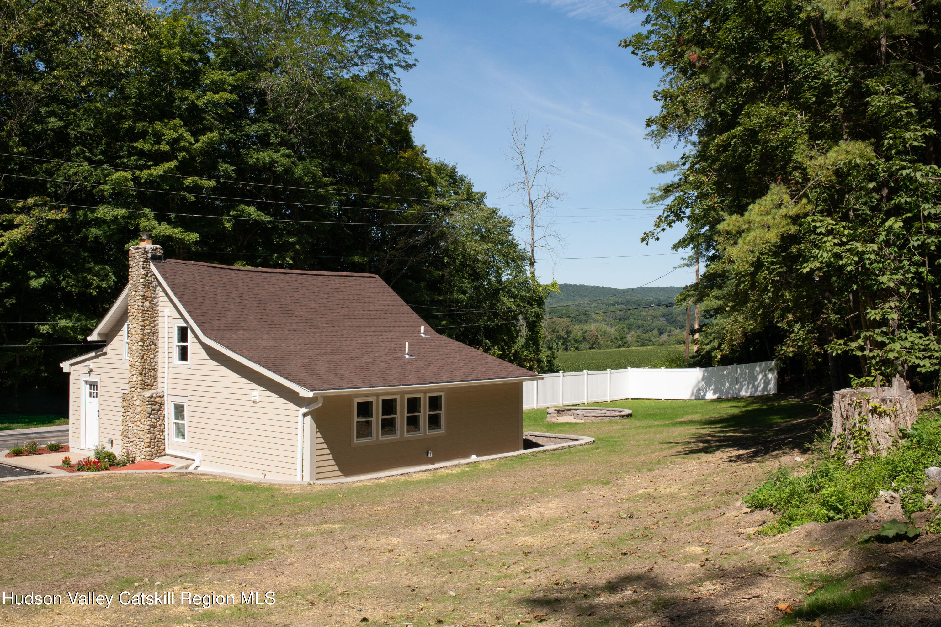 Beige house with a stone chimney, white fence, and fire pit in a wooded setting.
