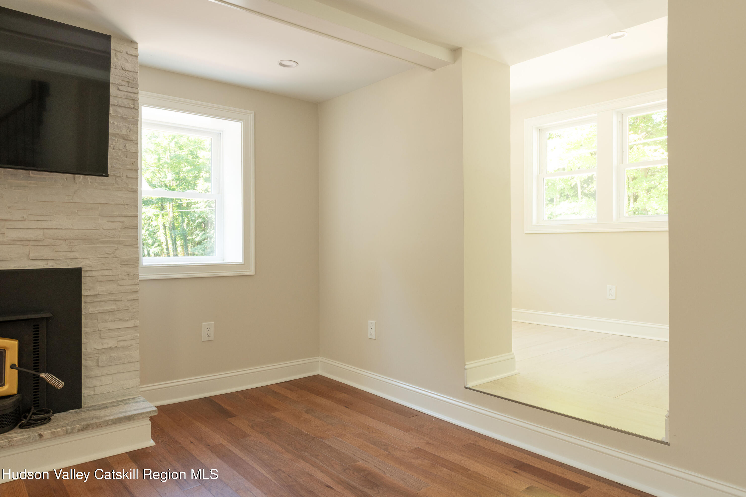 Living room with hardwood floors, a stone fireplace and television beside a bright adjoining room.