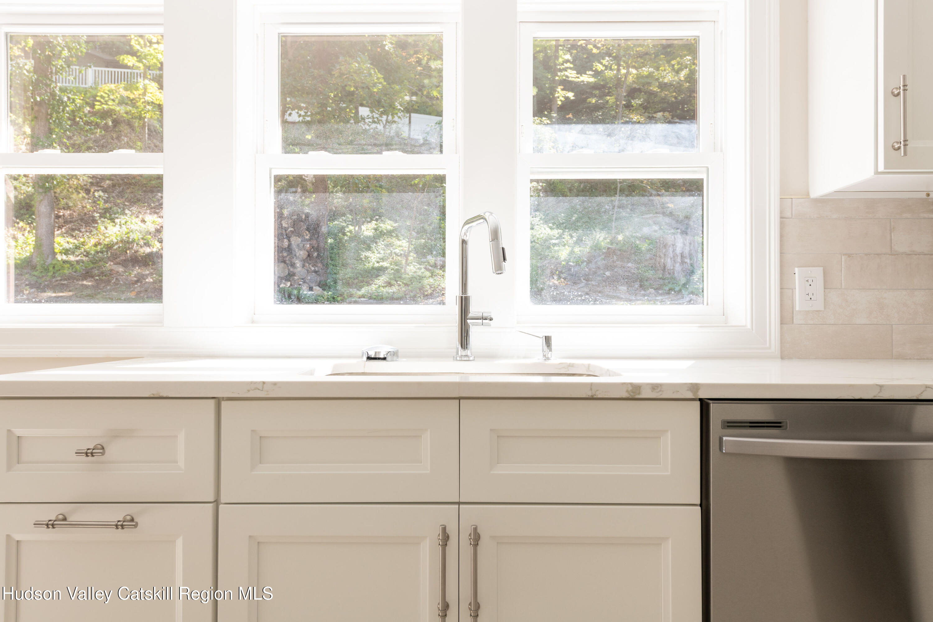 Kitchen sink beneath large windows, with white shaker cabinets and a stainless dishwasher.