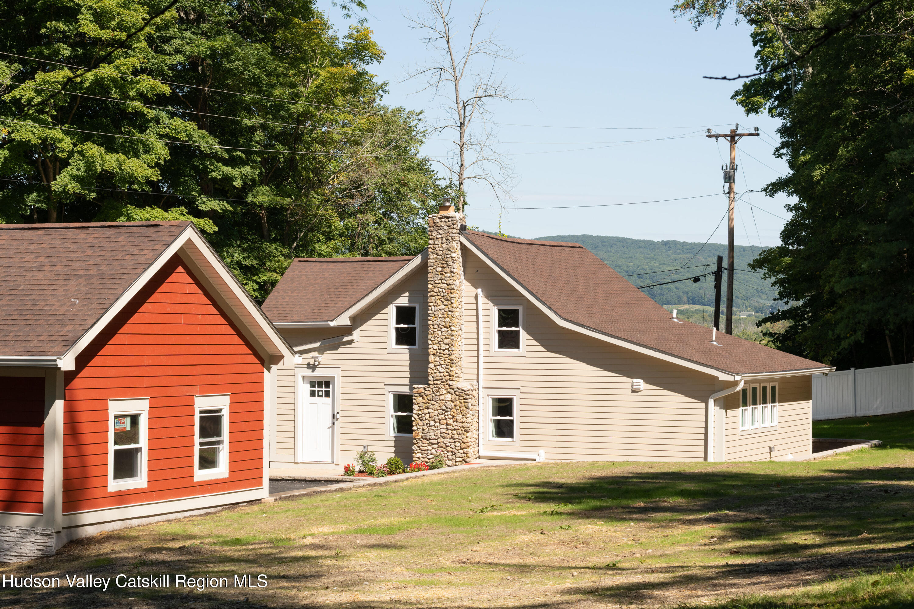 Beige home with a tall stone chimney and red front gable, backed by wooded hills.