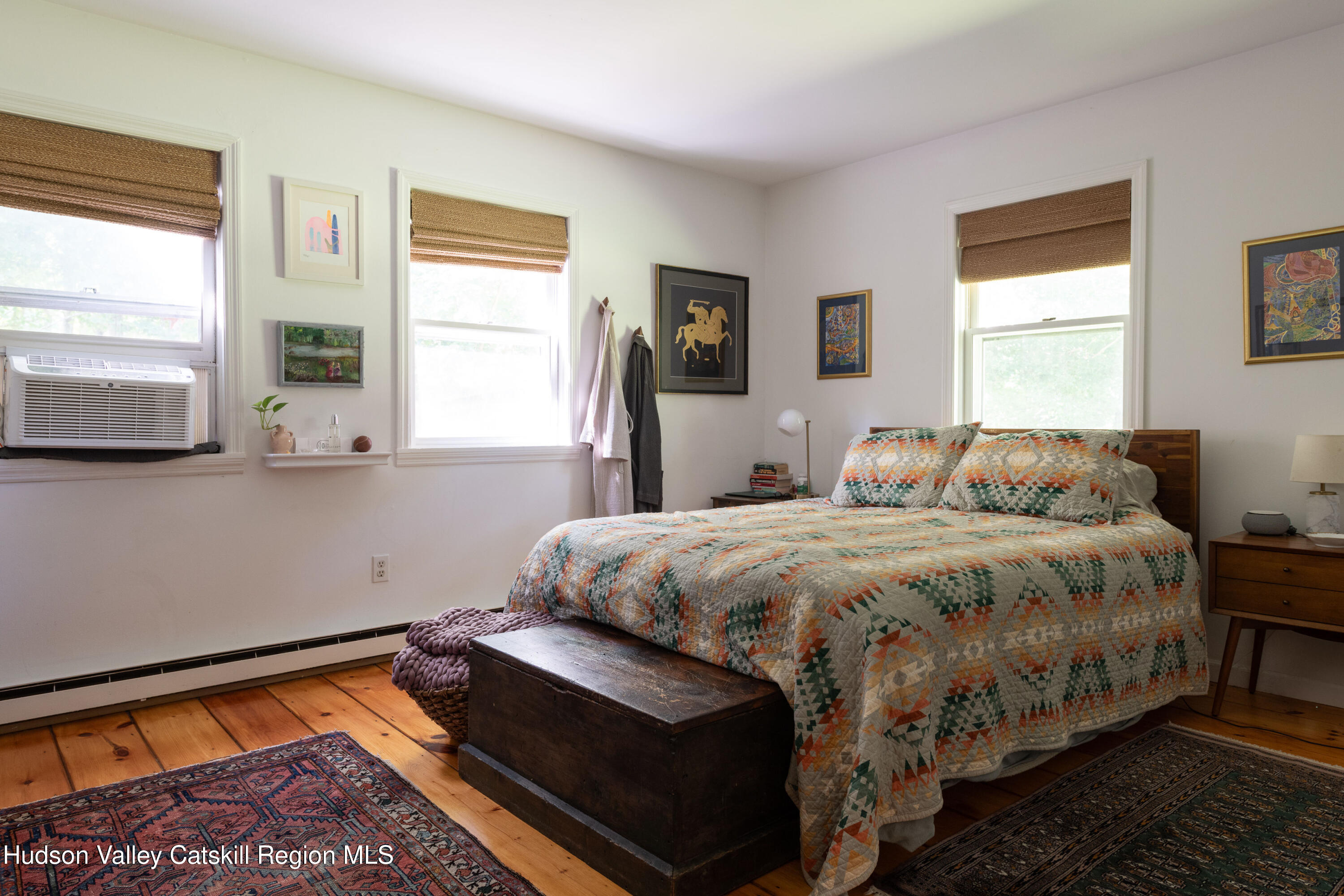 Bedroom with a geometric quilt, wooden chest, patterned rugs, and windows with woven shades.