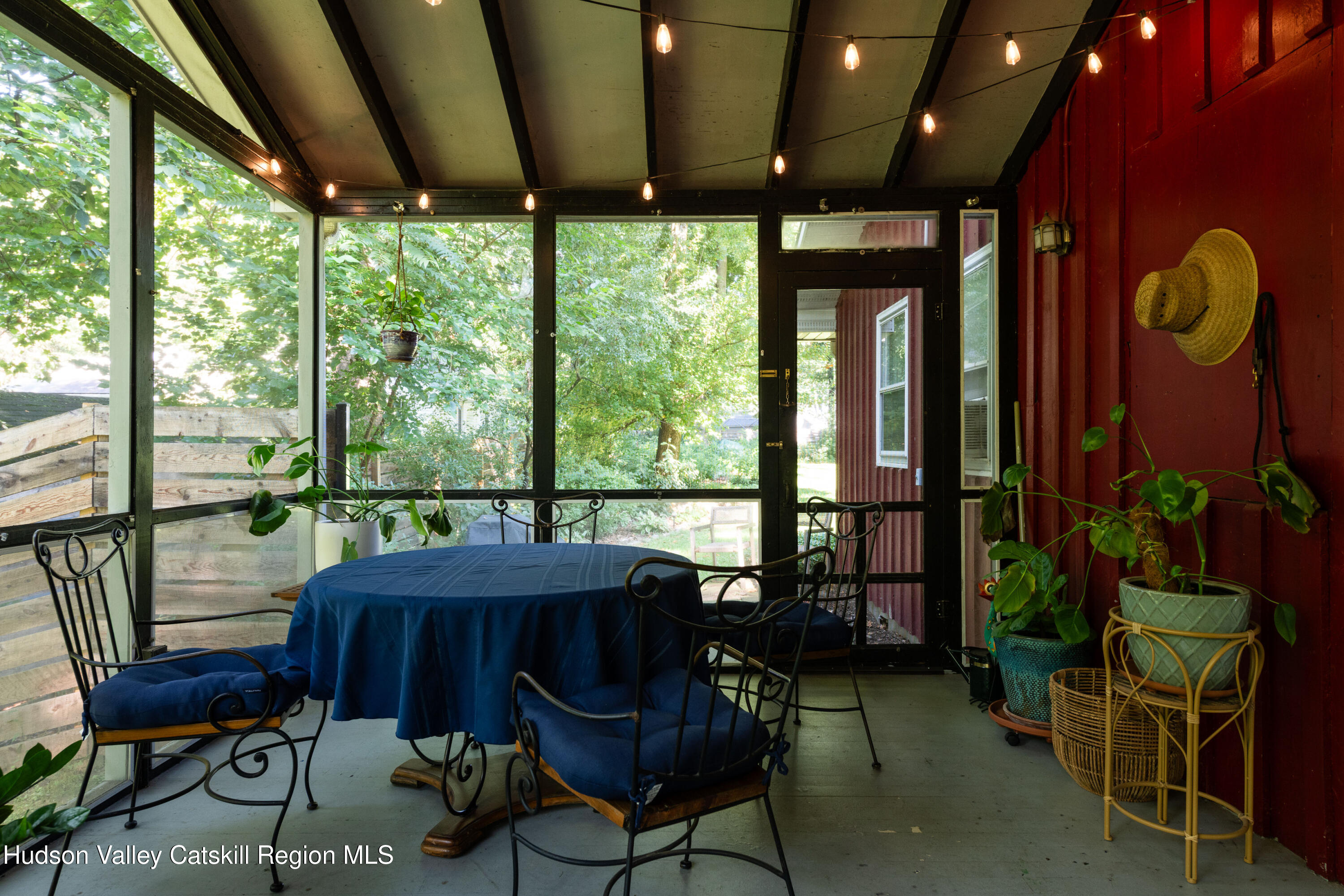 Enclosed porch with a blue-covered table, metal chairs, leafy views, and plants beside a red-paneled wall.