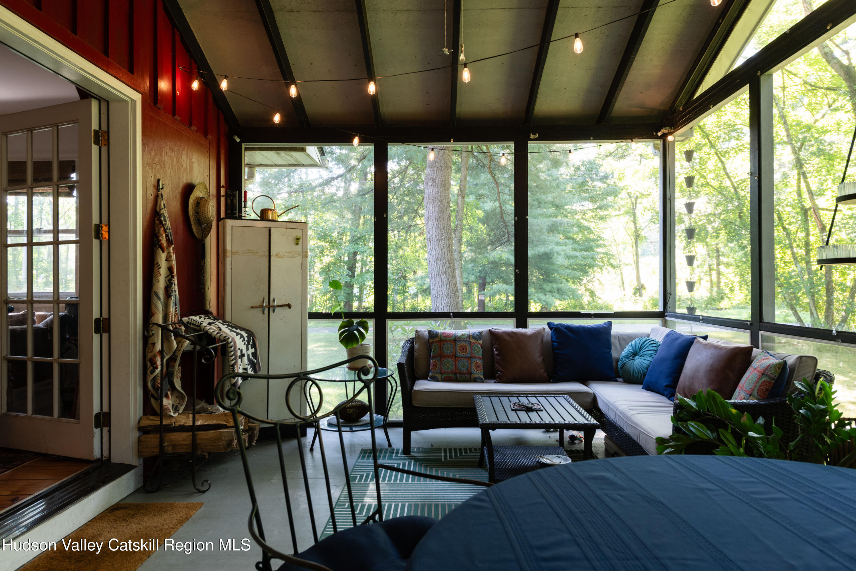 Screened porch with a cushioned wicker sectional, string lights, and large windows overlooking trees.