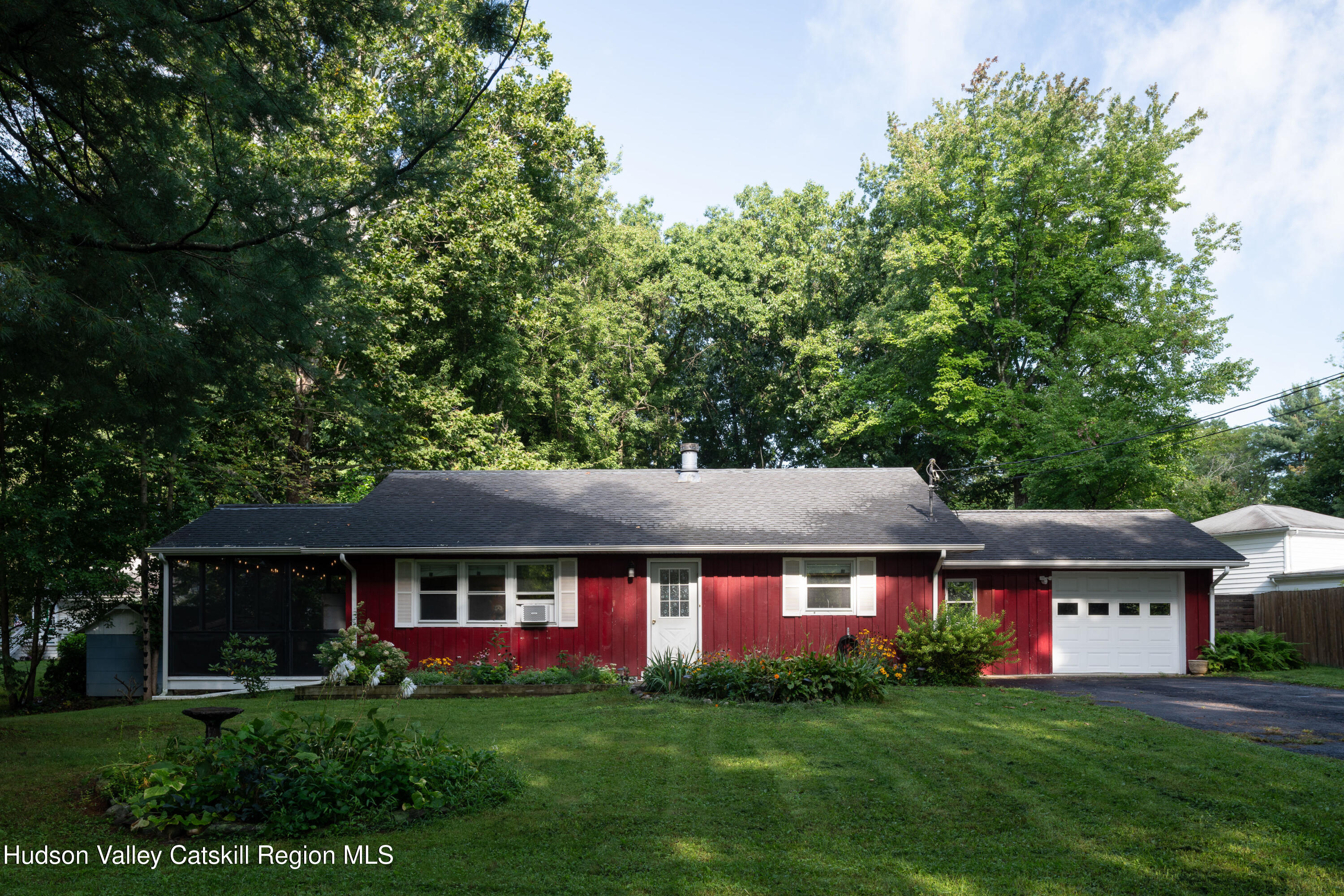 Red single-story house with a screened porch, attached garage, and flower beds beneath mature trees.