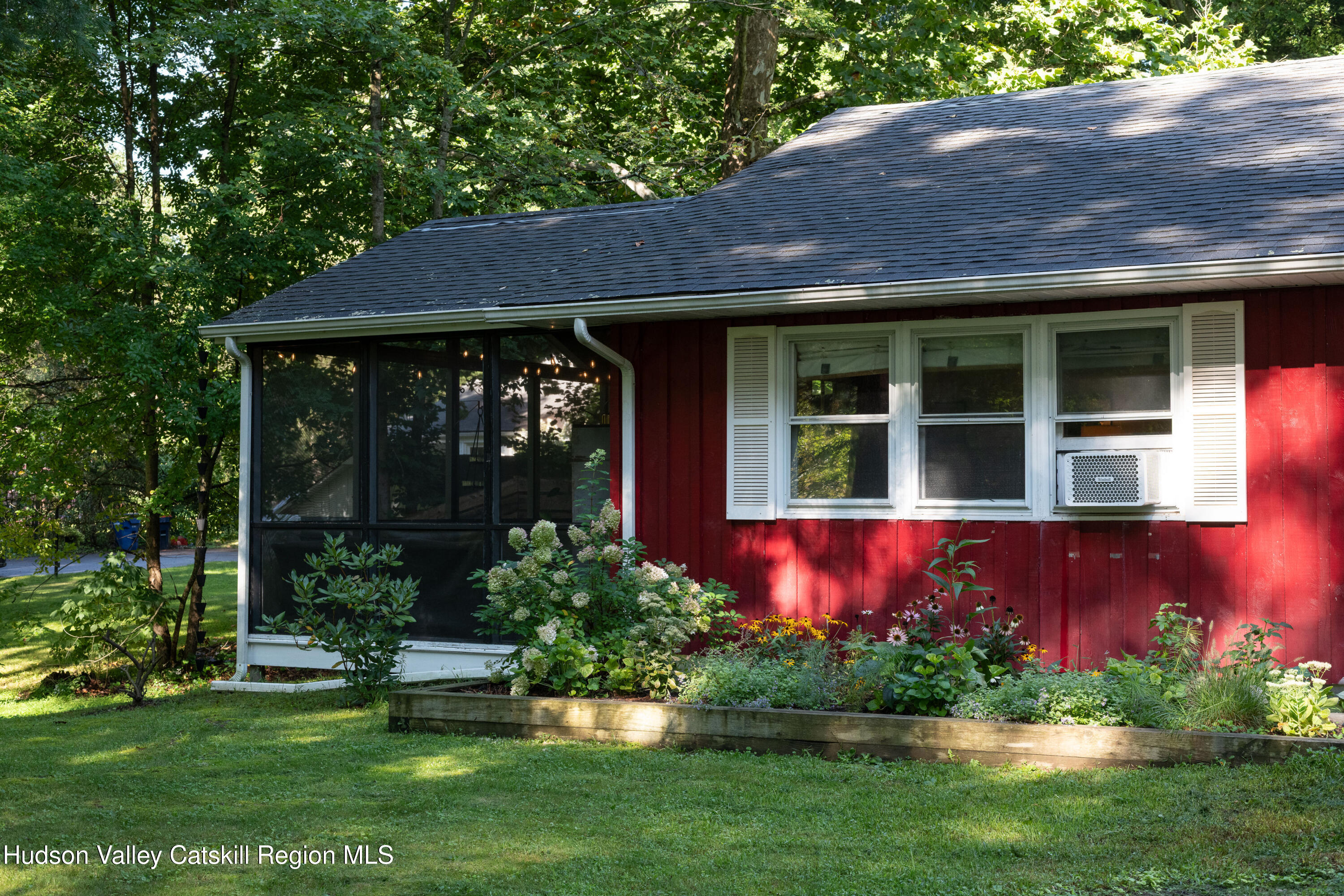 Red-sided home with a screened porch and colorful flower garden among trees.