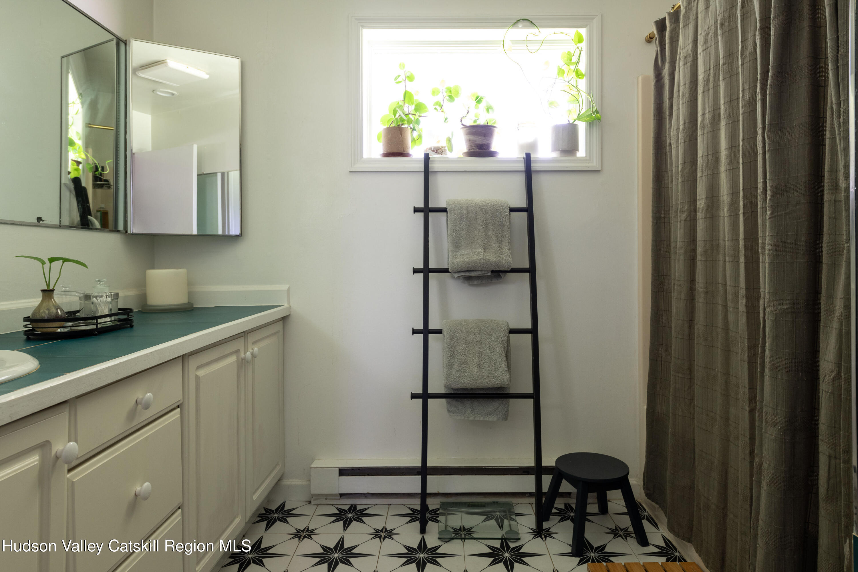 Bathroom with teal vanity, plant-filled window, towel ladder, and geometric black-and-white floor tiles.