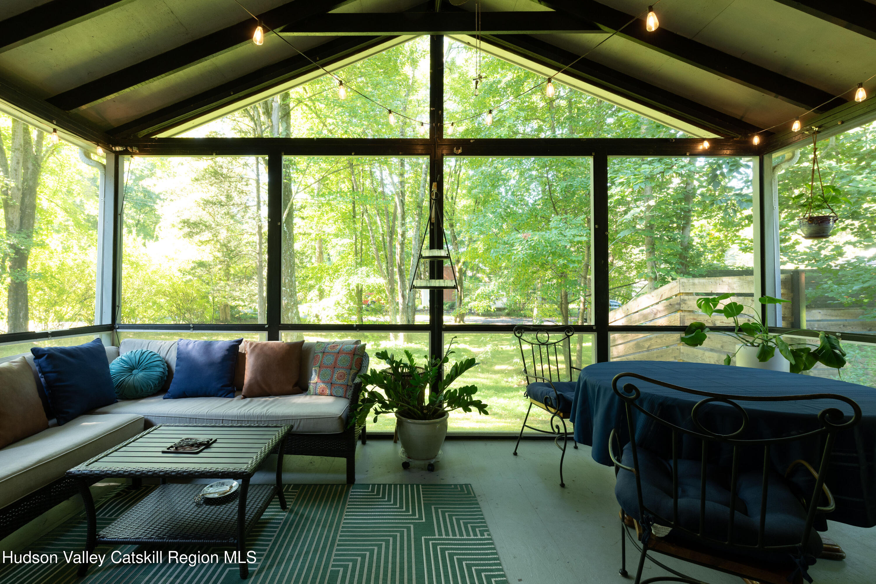 Furnished glass-enclosed sunroom with a sectional sofa, dining table, and leafy yard views.