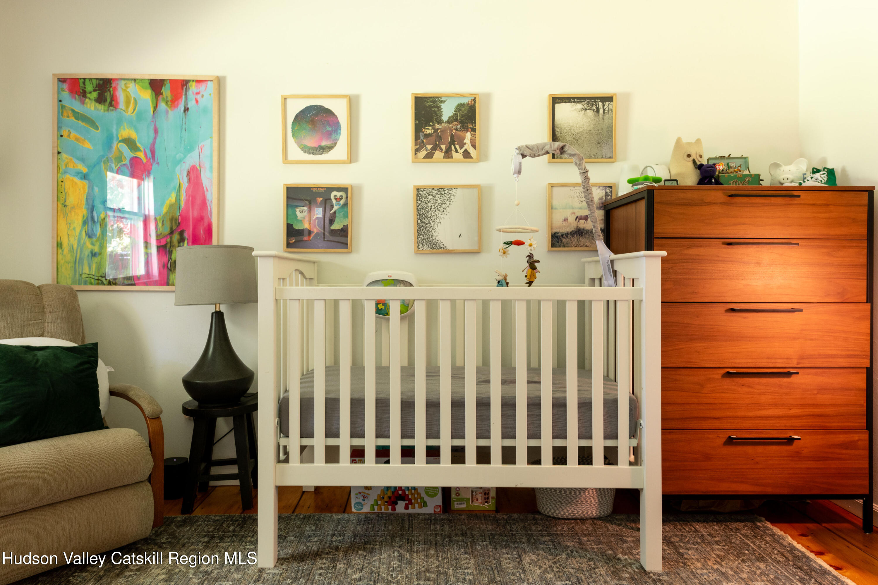 Nursery with a white crib, wooden dresser, colorful wall art, and an upholstered armchair.