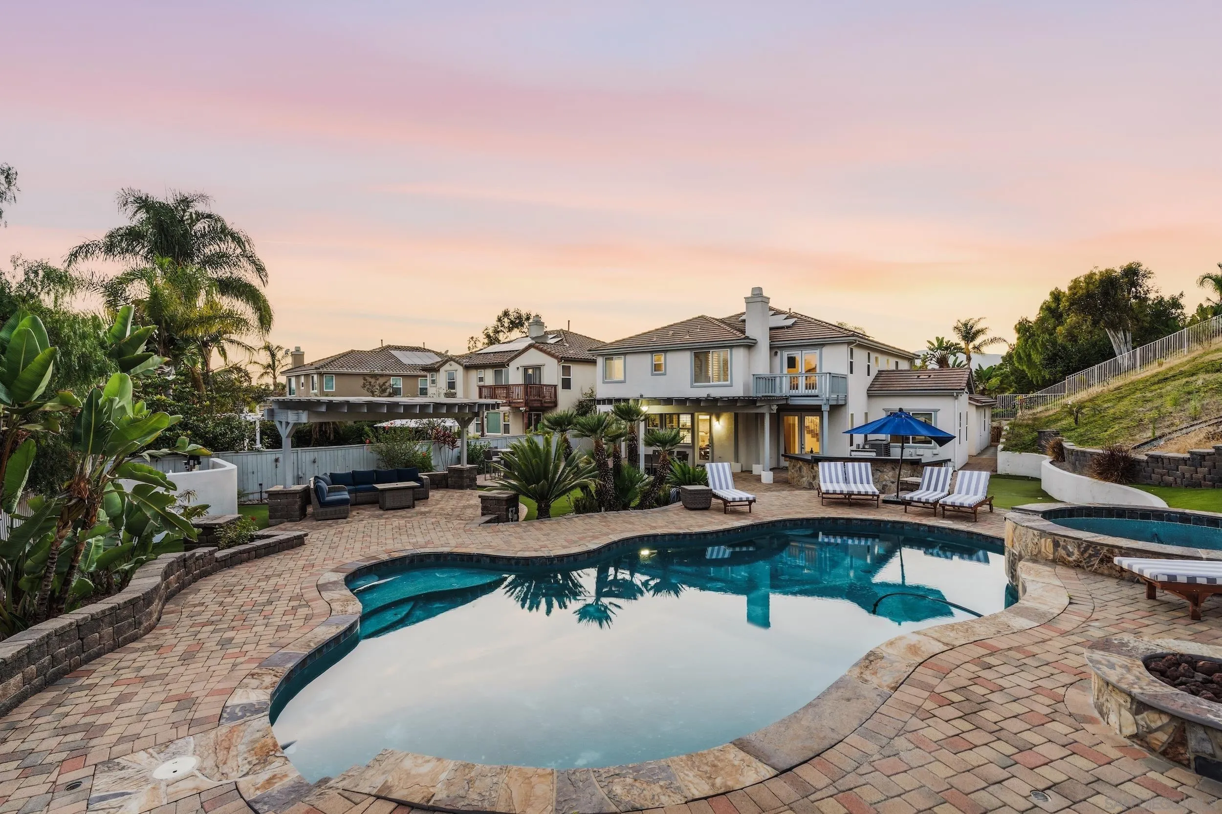 Two-story home behind a curved pool and stone patio with outdoor seating and tropical landscaping at dusk.