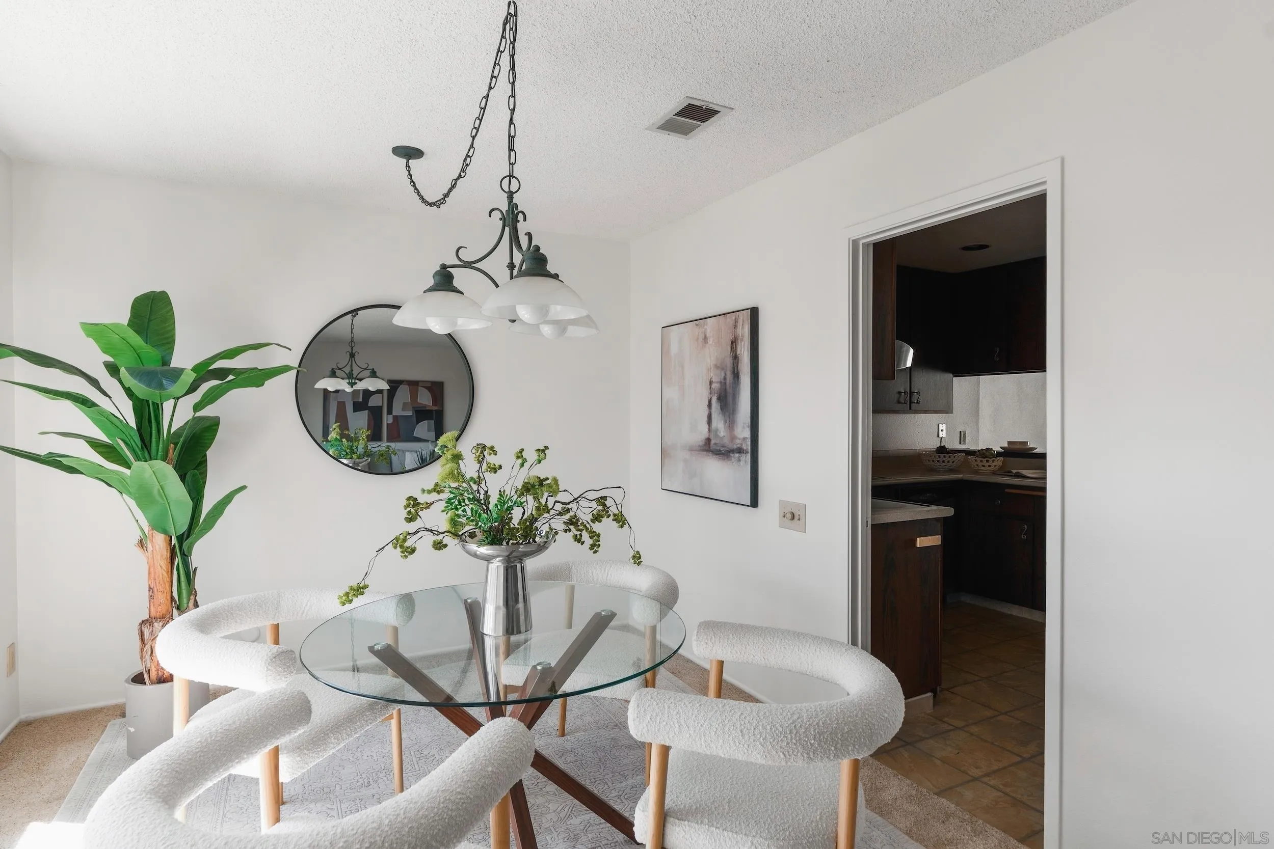 Dining area with a glass table, white boucle chairs, chandelier, and doorway to the kitchen.