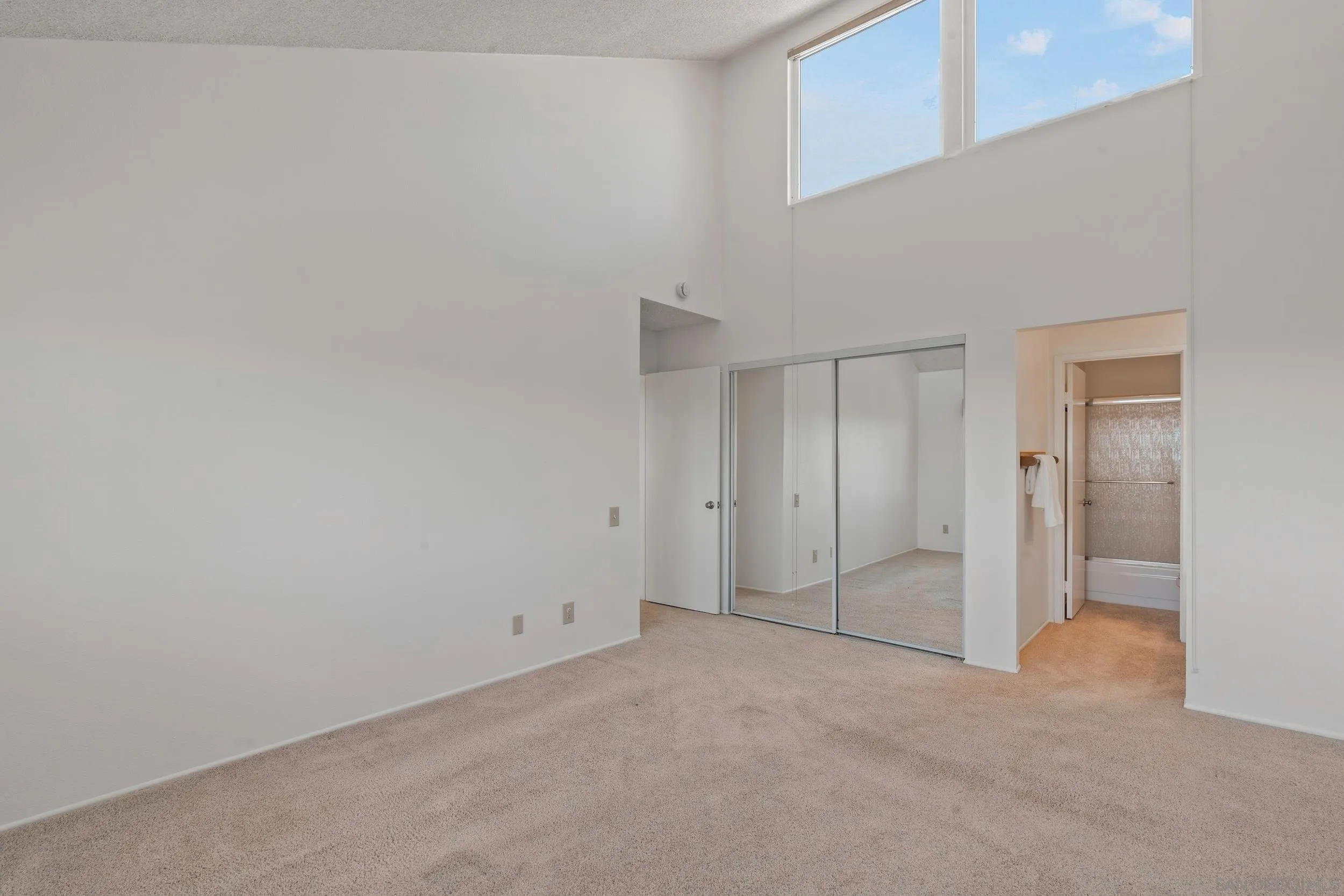 Empty carpeted bedroom with high windows, mirrored closet doors, and an adjoining bathroom.