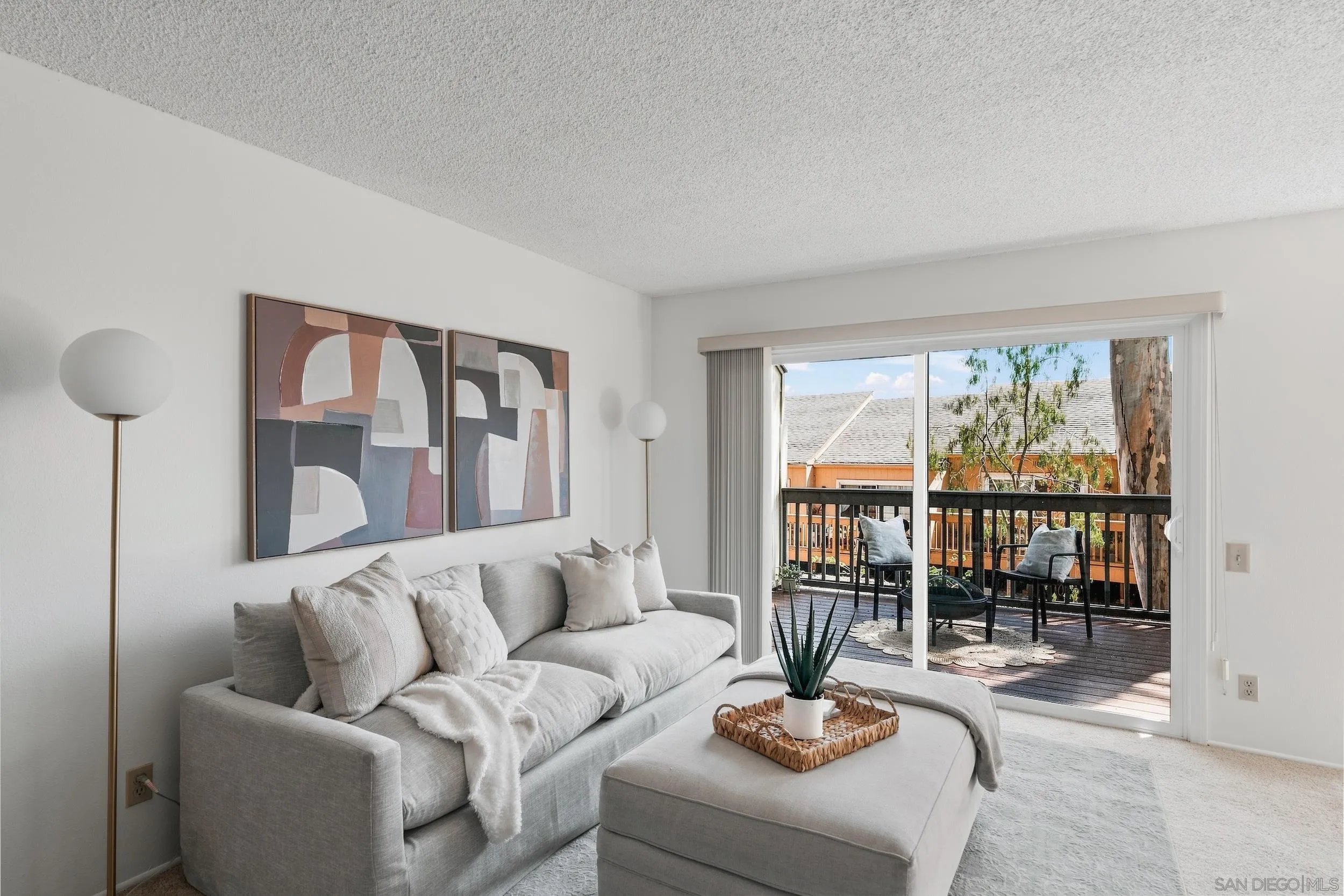 Living room with a light gray sofa and abstract art, opening onto a balcony with two chairs.