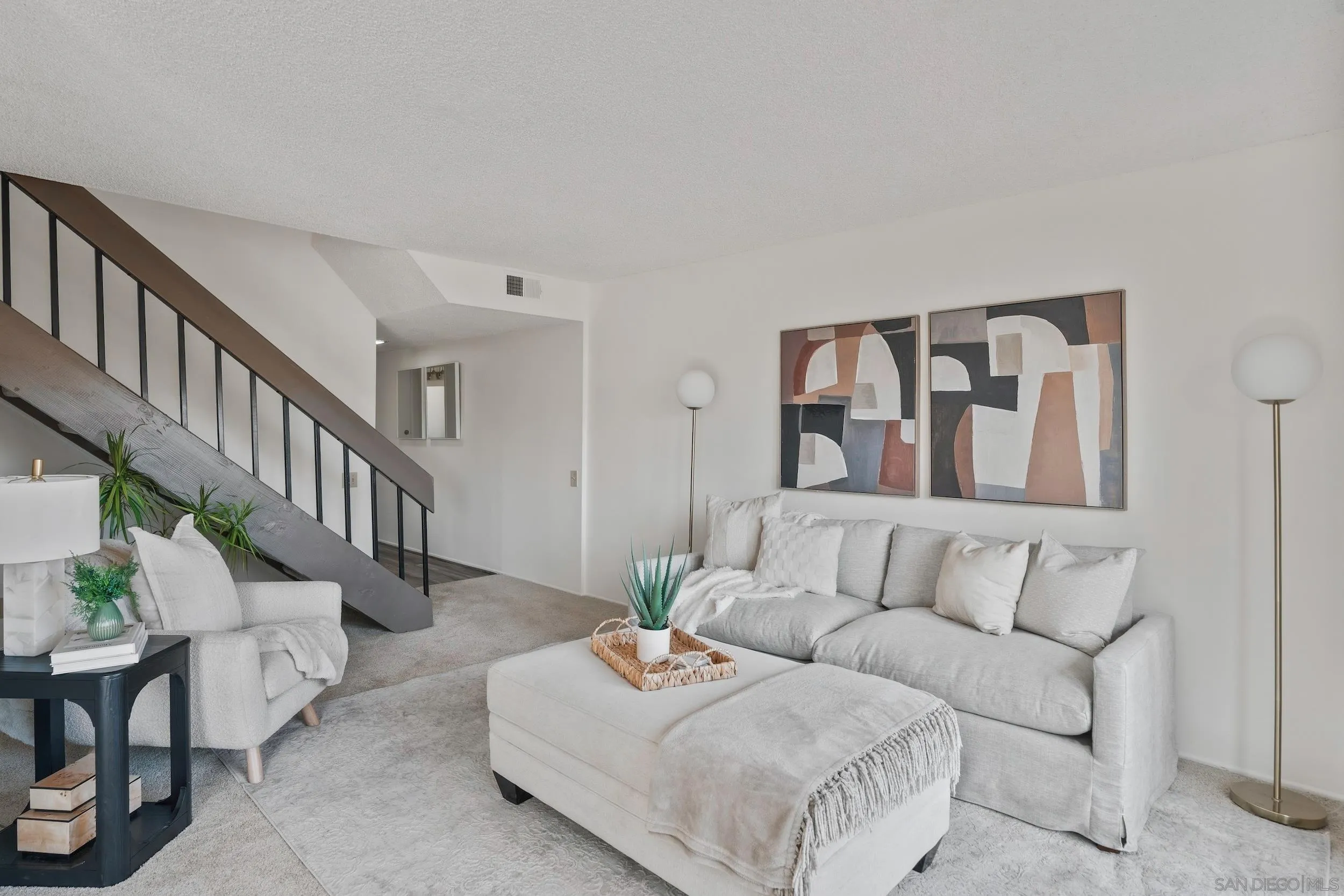 Staged living room with a gray sofa, oversized ottoman, abstract art, and dark metal staircase.